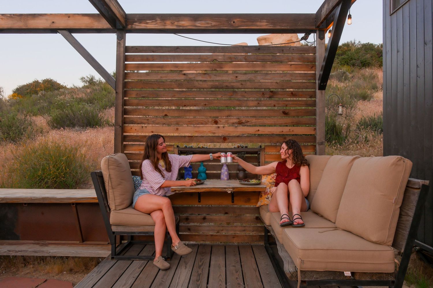 Two women toasting drinks on a rustic wooden deck patio with outdoor seating at sunset in a desert landscape.