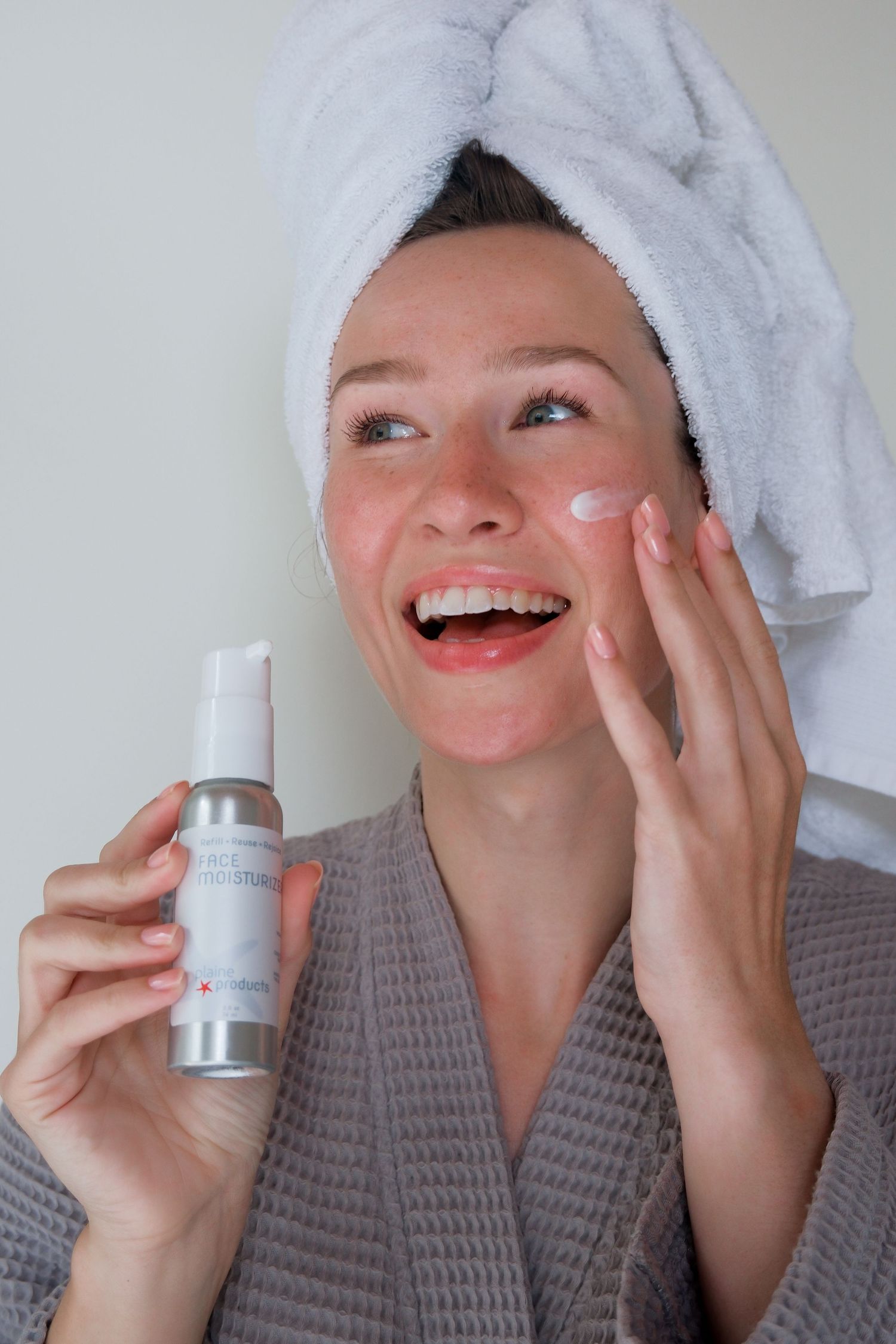 Smiling woman in robe and towel turban applying face moisturizer from silver pump bottle after shower.
