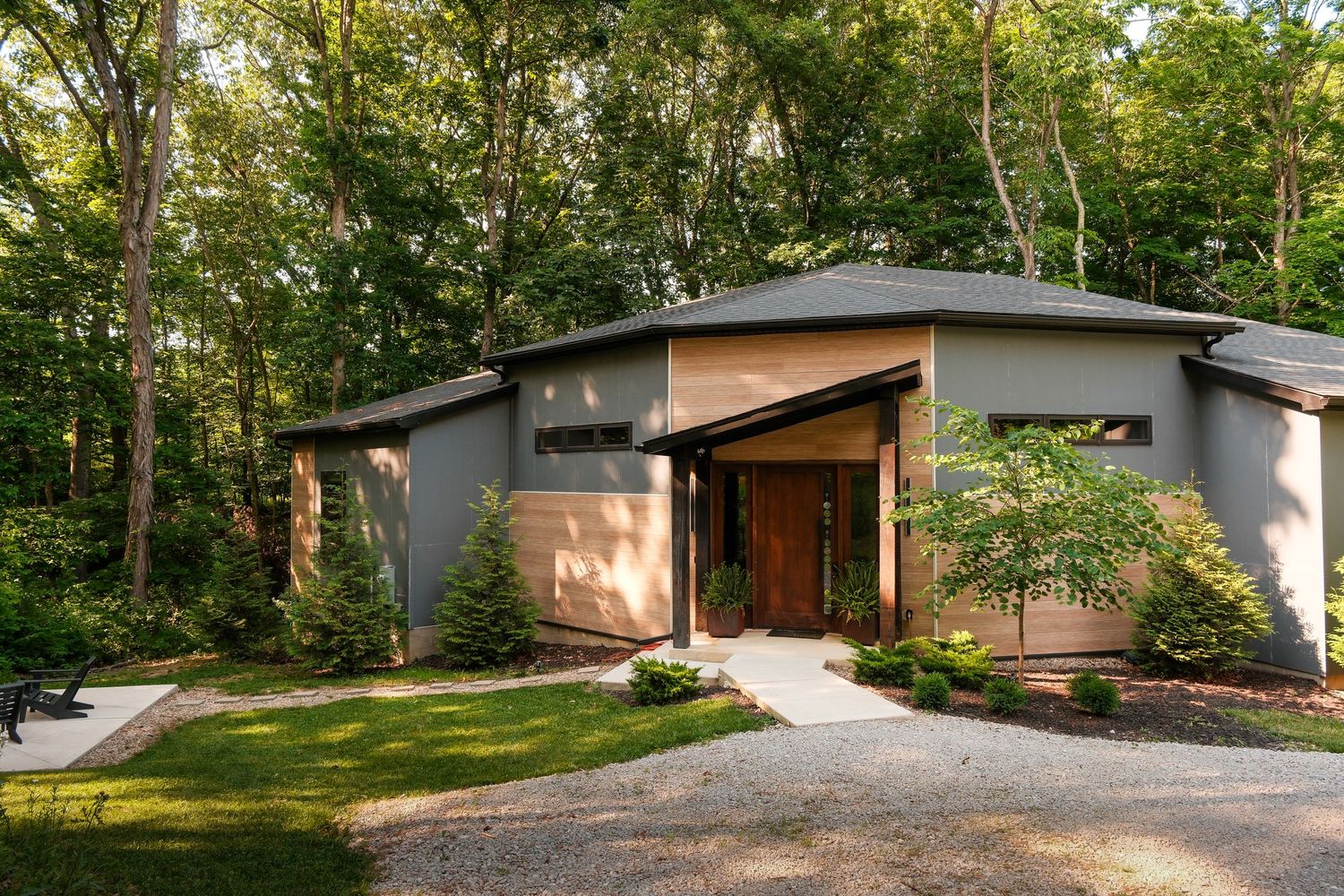 Modern mid-century home with gray exterior, wood-accented entry, and lush surrounding trees on a gravel driveway.
