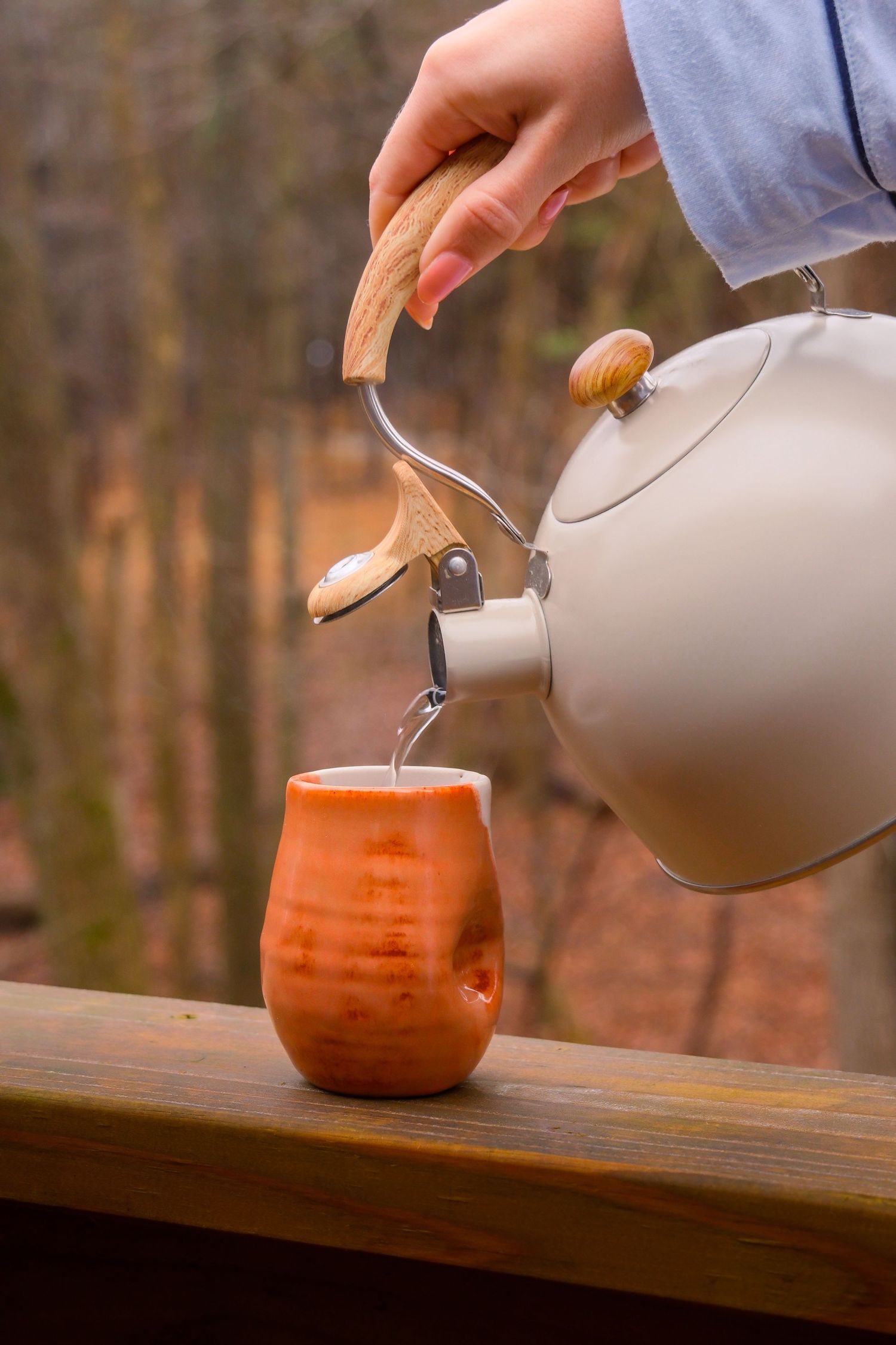 Hand pouring hot water from a white kettle into an orange ceramic mug on a wooden railing outdoors.
