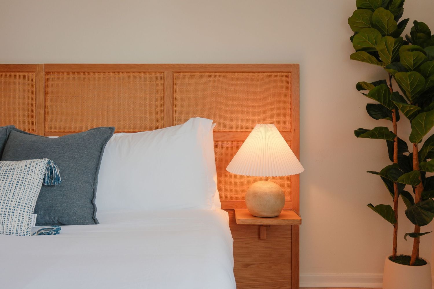 Cozy bedroom with wooden headboard, white pillows, glowing table lamp, and a fiddle leaf fig plant in warm light.