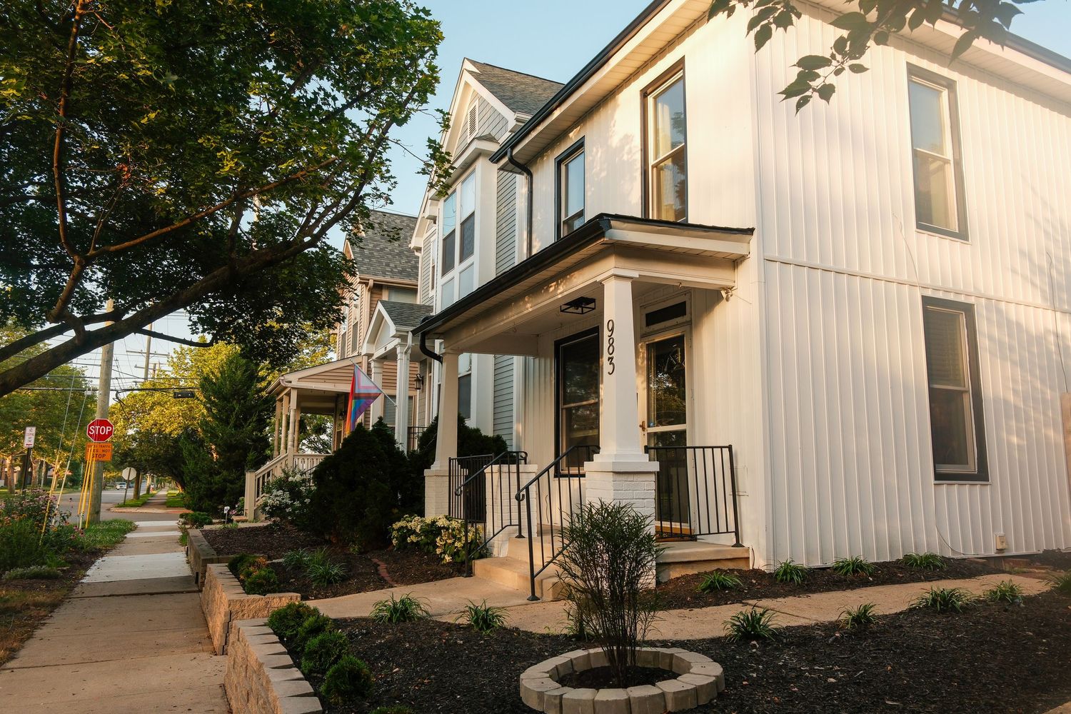 White two-story colonial home with covered porch, landscaped garden, and tree-lined sidewalk in a residential neighborhood.