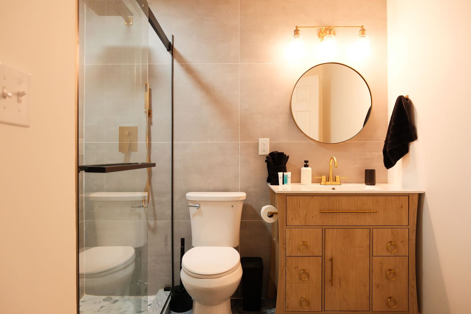Modern bathroom with walk-in glass shower, wood vanity, round mirror, and gold fixtures on beige tile walls.