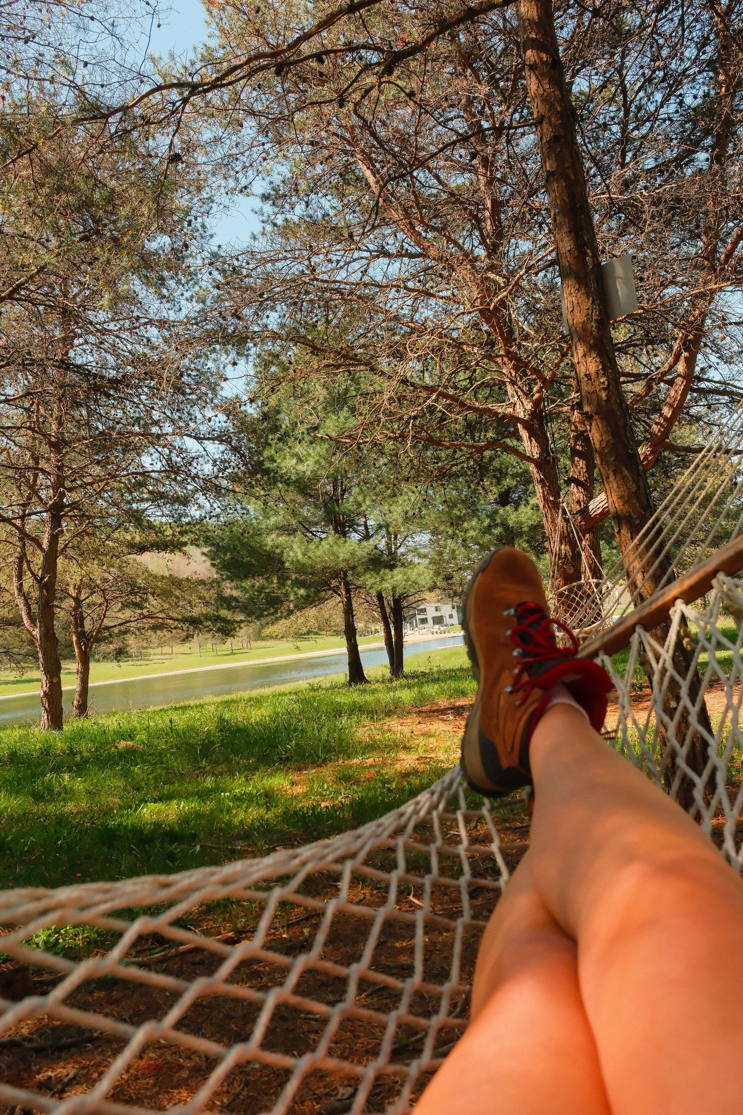 Person relaxing in a rope hammock outdoors, legs stretched out wearing brown hiking boots, surrounded by trees on a sunny day.