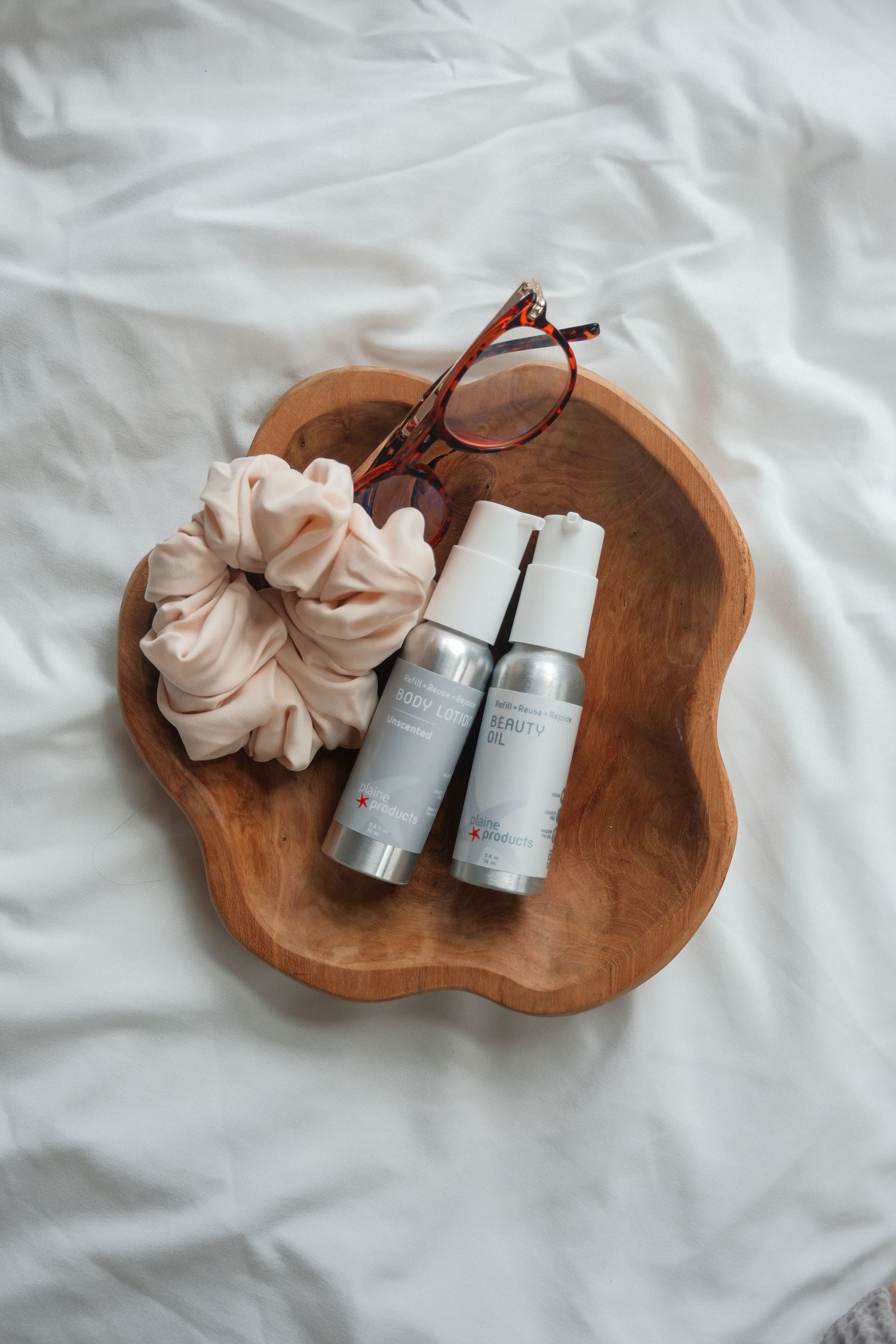 Wooden tray on white bedding with two skincare spray bottles, a pink scrunchie, and red tortoiseshell glasses.