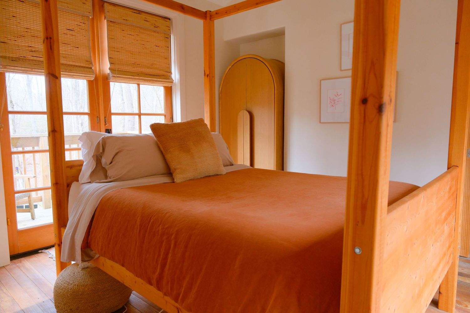 Cozy bedroom with orange four-poster bed, warm wood accents, arched mirror, and natural light from large windows.