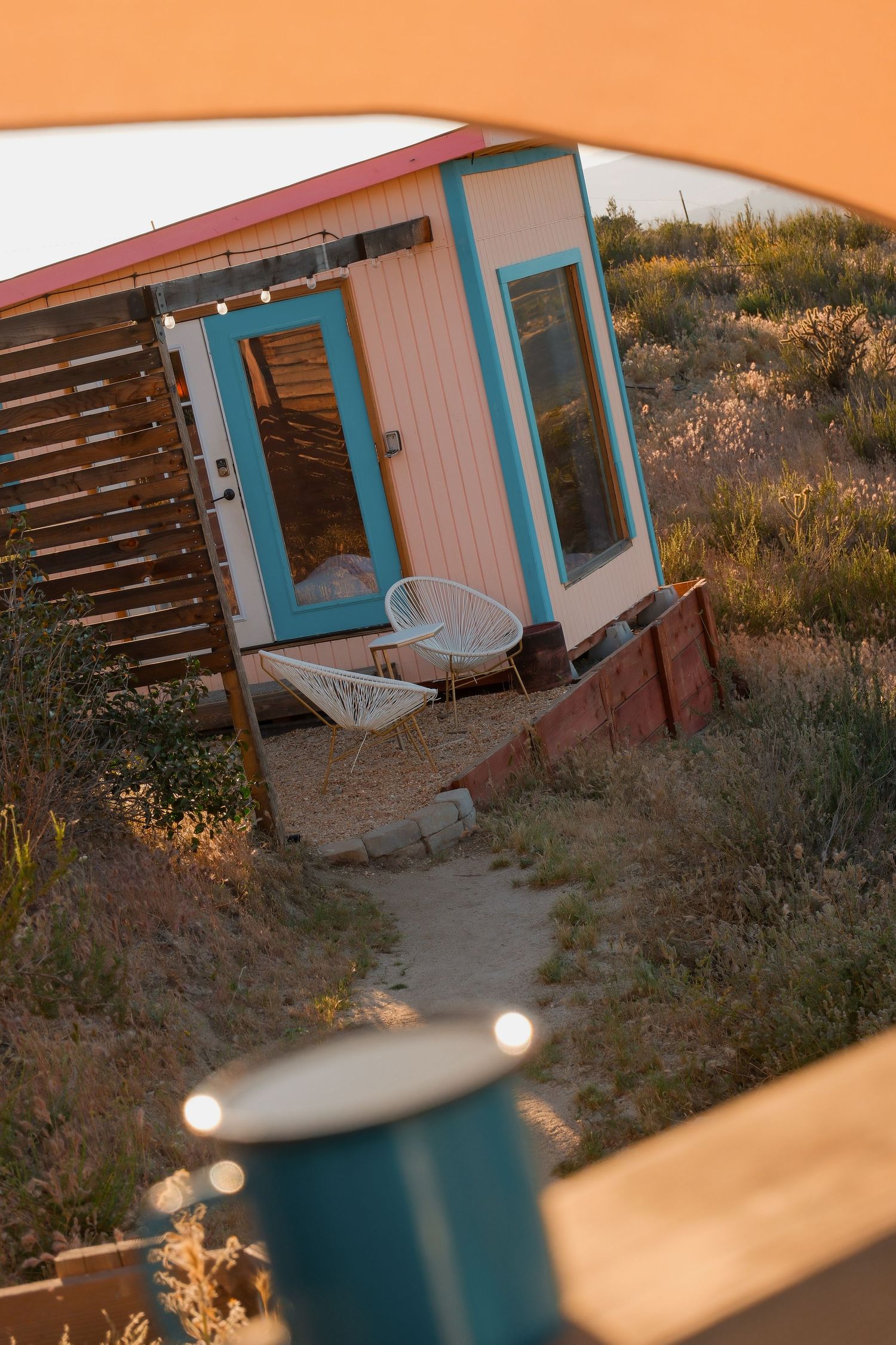 Tiny house with turquoise door and white chairs nestled in desert landscape at golden hour, viewed through tent opening.