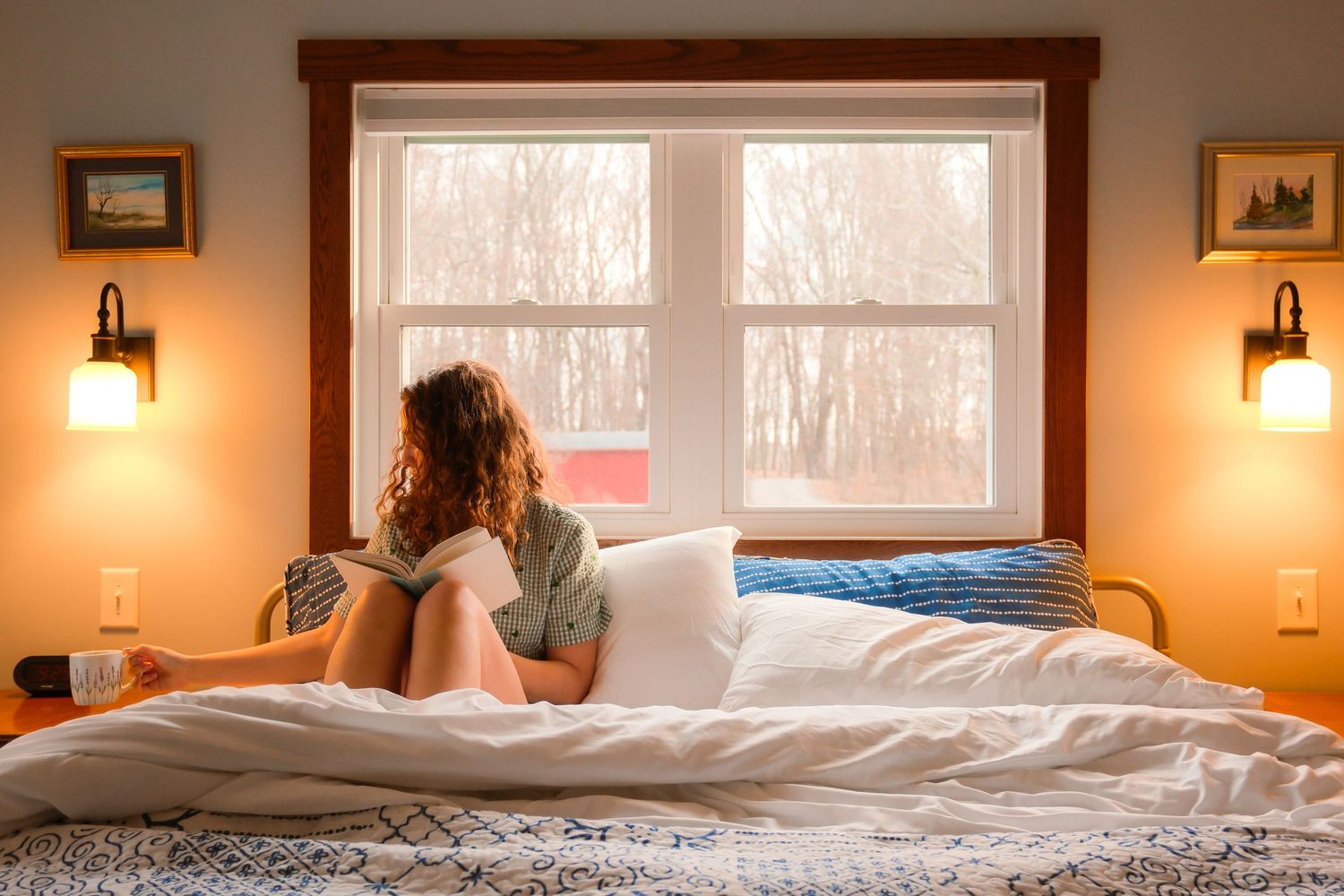 A cozy bedroom with warm lighting features multiple windows, wall sconces, and a comfortable bed with white linens.