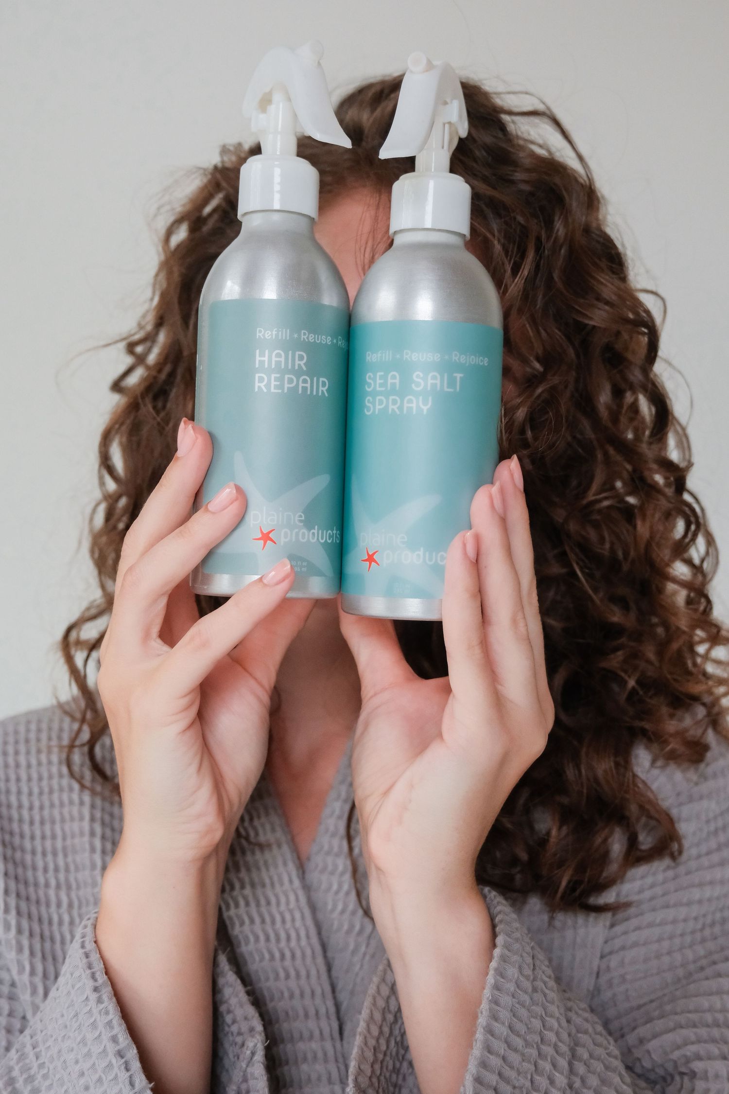 Woman with curly hair holding Plain Products Hair Repair and Sea Salt Spray bottles in teal packaging.