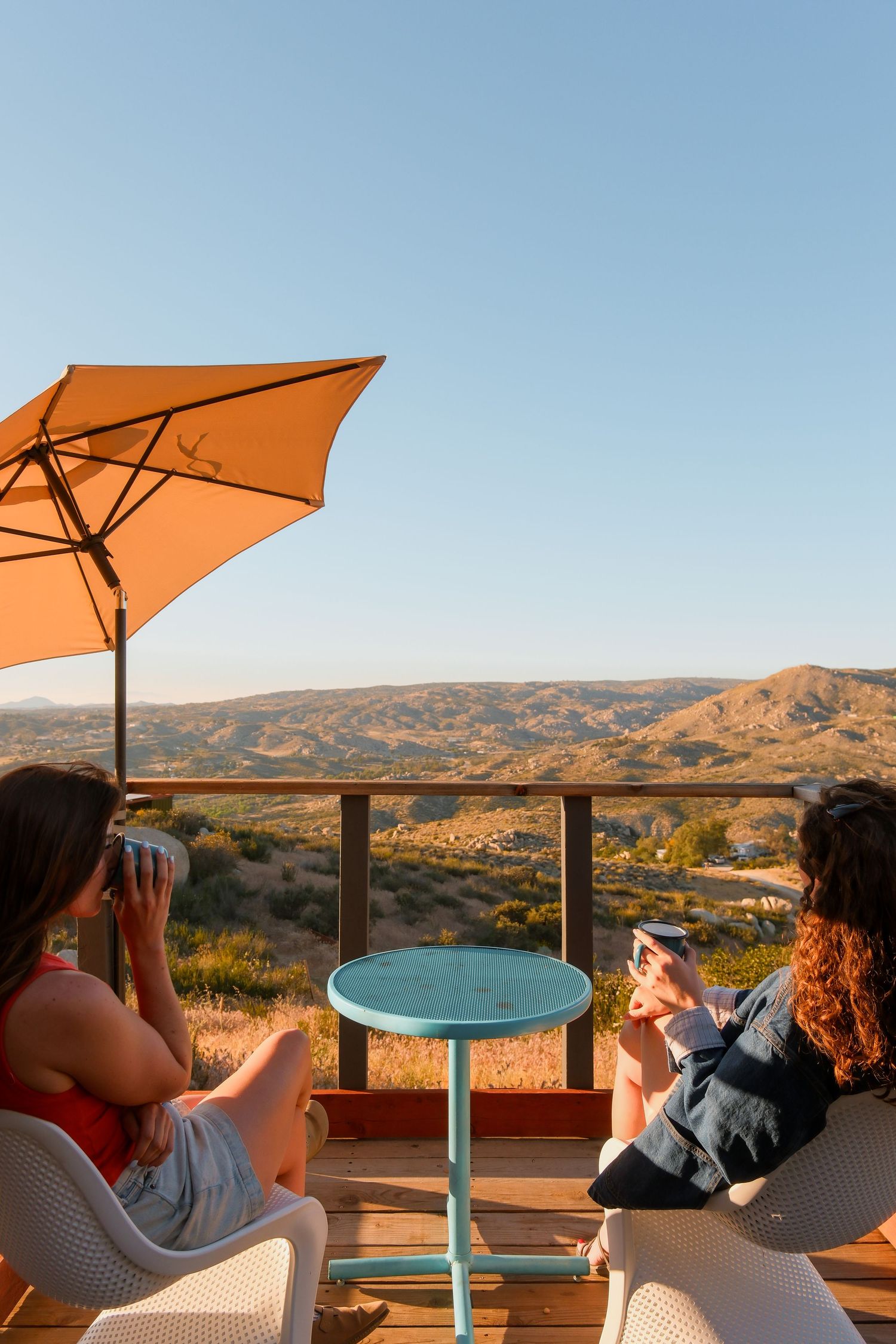 Two women sitting on a deck with an orange umbrella enjoying drinks and mountain views at golden hour.