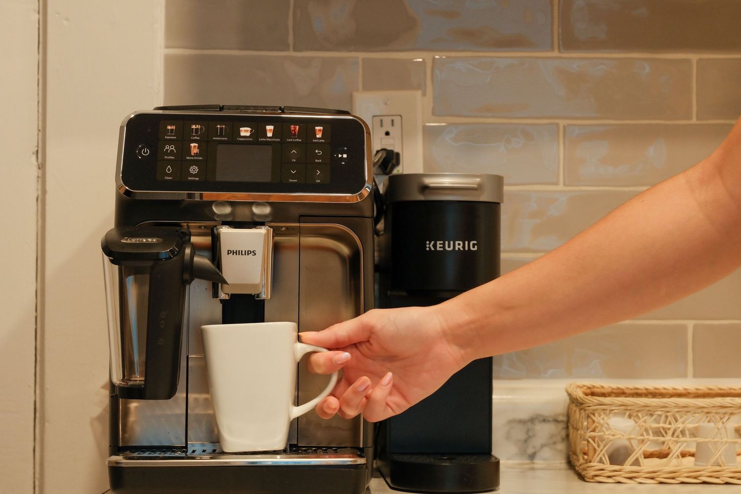 A hand reaching for a white coffee mug on a Philips espresso machine next to a Keurig milk frother.