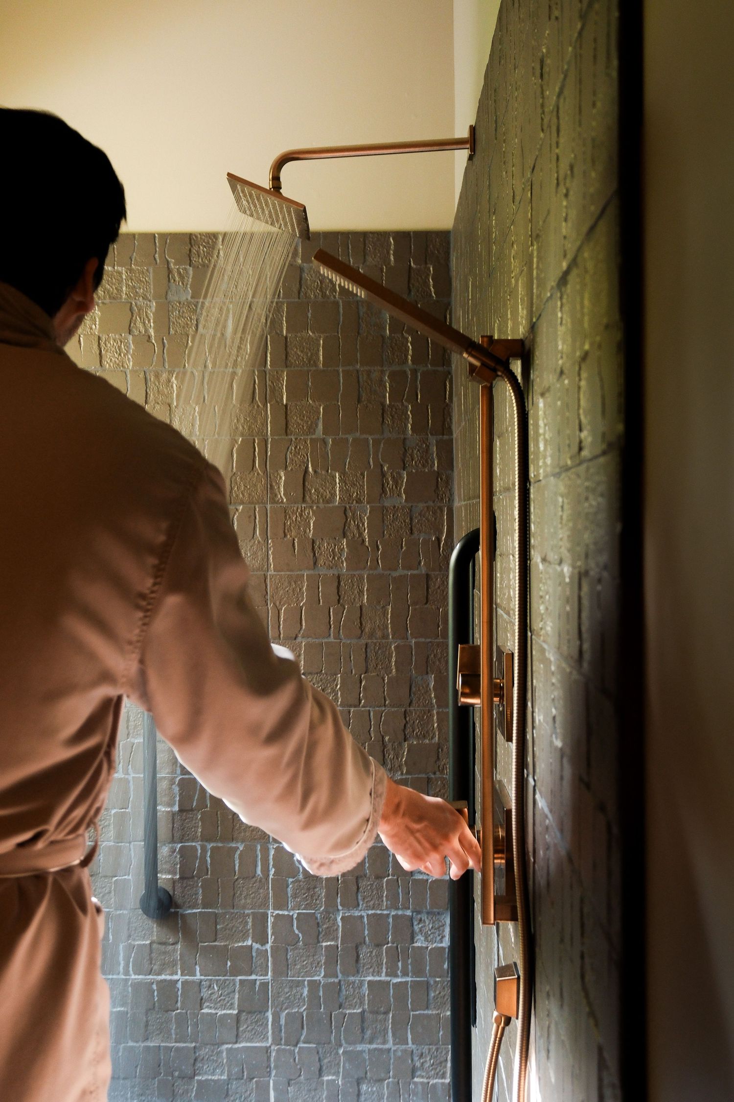 Person in bathrobe adjusting copper rainfall shower system in a modern stone-tiled bathroom.