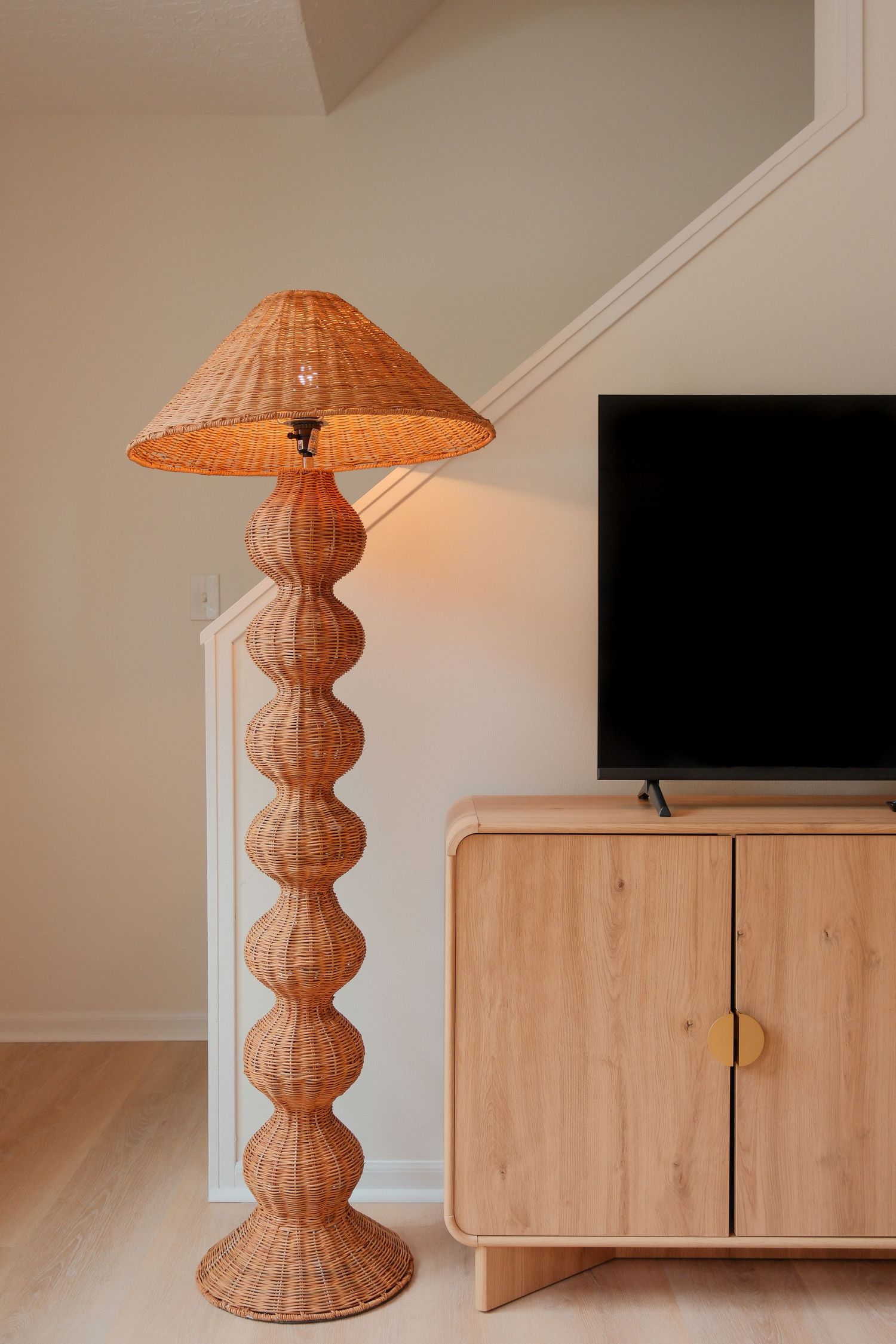 Tall wicker rattan floor lamp with stacked ball base beside a light wood TV console with flat screen television.
