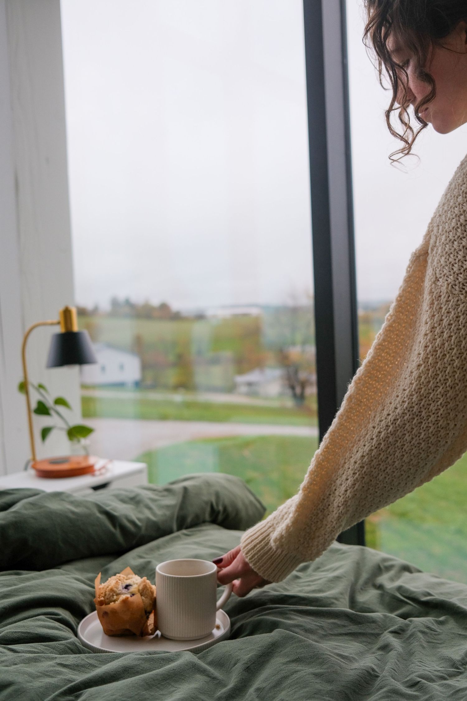 Person arranging breakfast tray with coffee and pastry on green bedding near window.