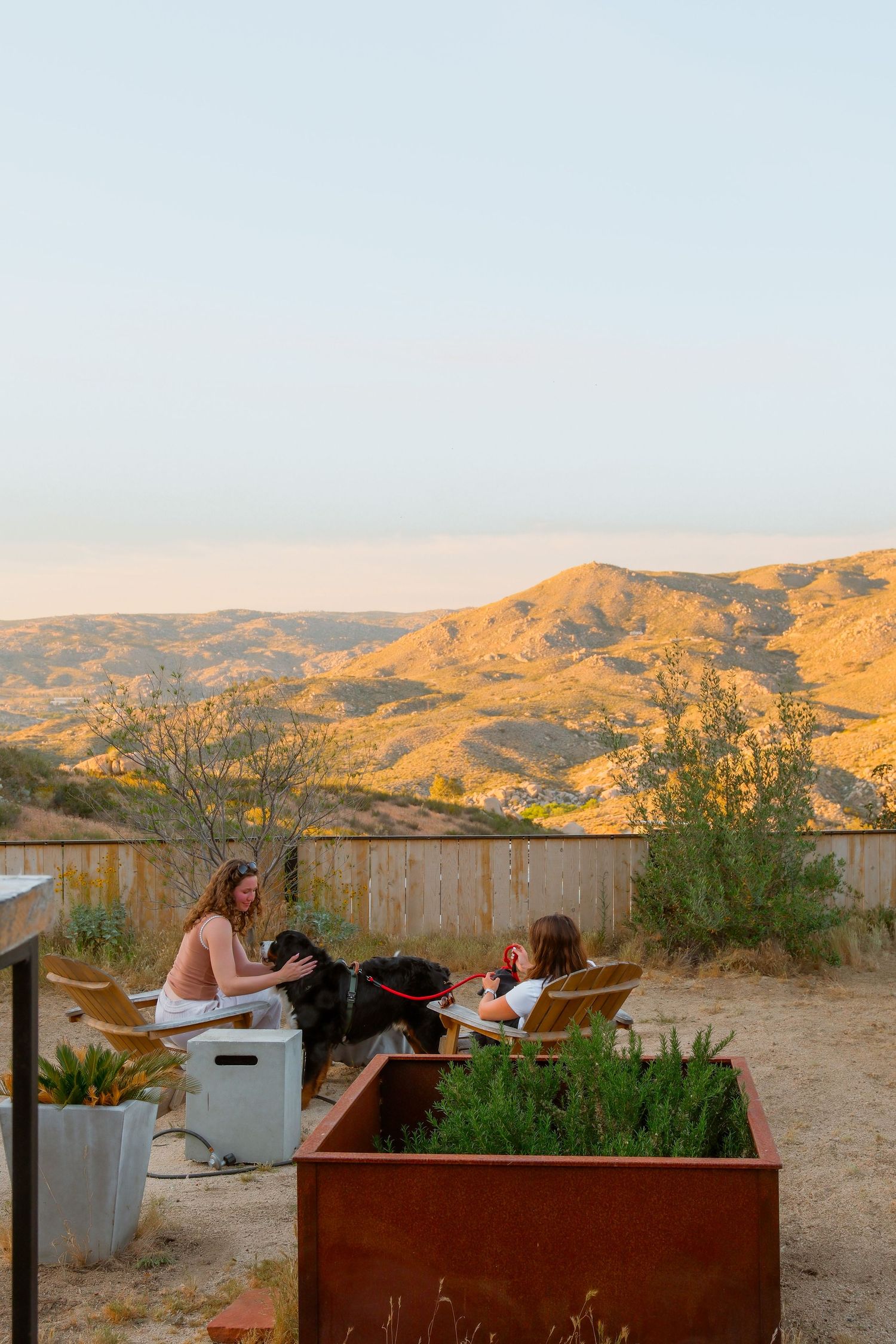 Two people relax outdoors with a black dog, enjoying golden hour views of rolling desert hills from a rustic patio.