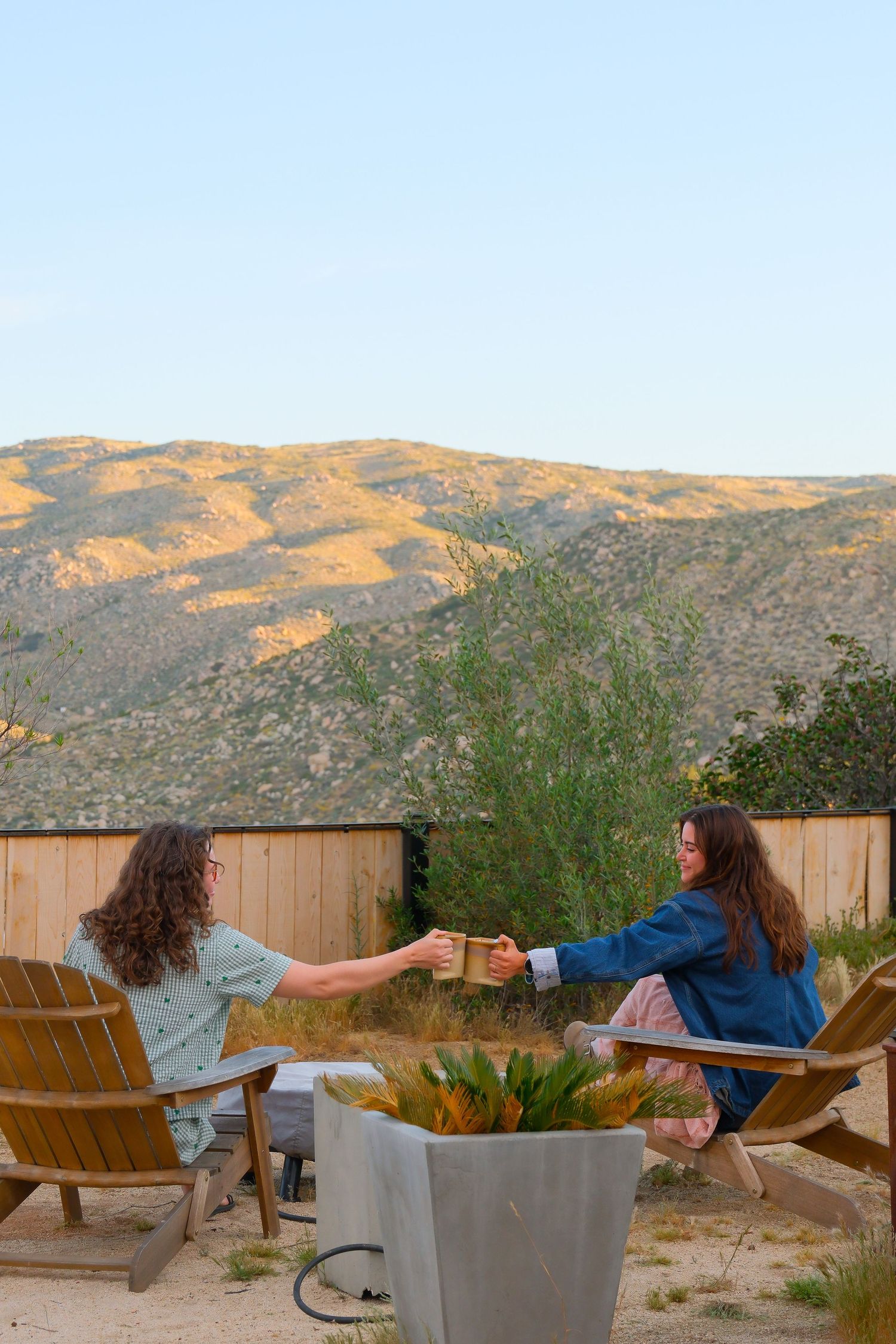 Two women toast drinks in Adirondack chairs by a fire pit with stunning golden mountain views at sunset.