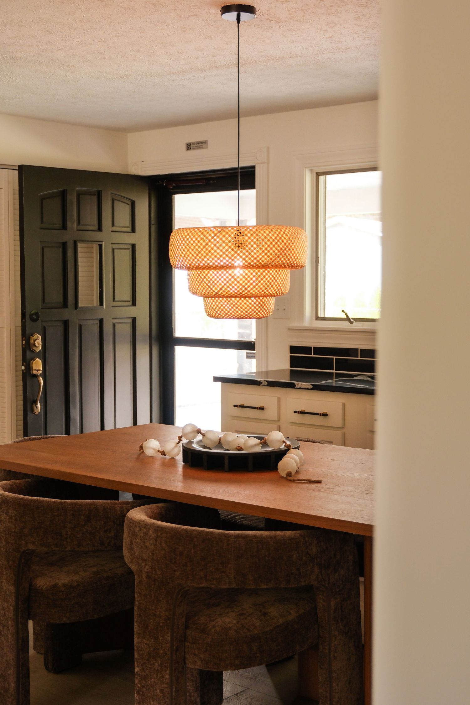 Dining area with wood table, brown chairs, woven pendant light, black door, and white kitchen in background.