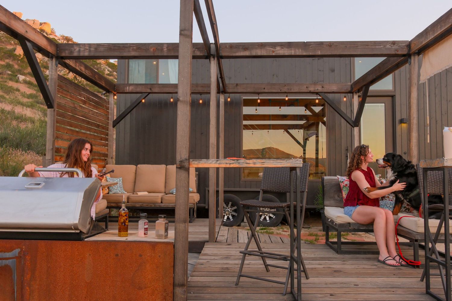 Two women relax on a rustic wooden deck with a hot tub and black dog at sunset near a modern cabin.