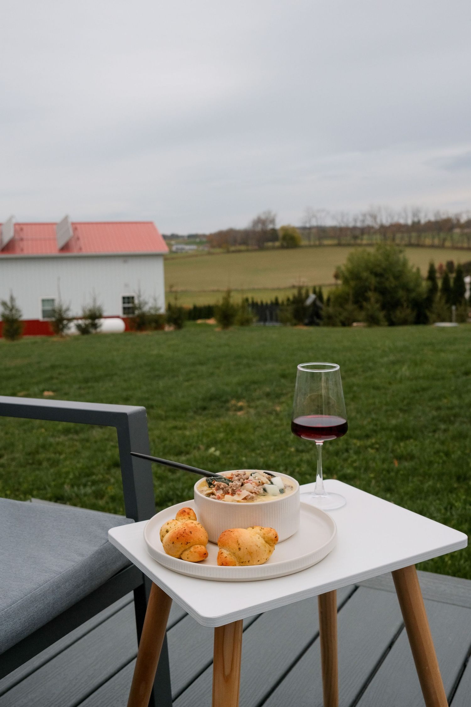Outdoor dining setup with wine and food on small white tables overlooking a rural landscape.