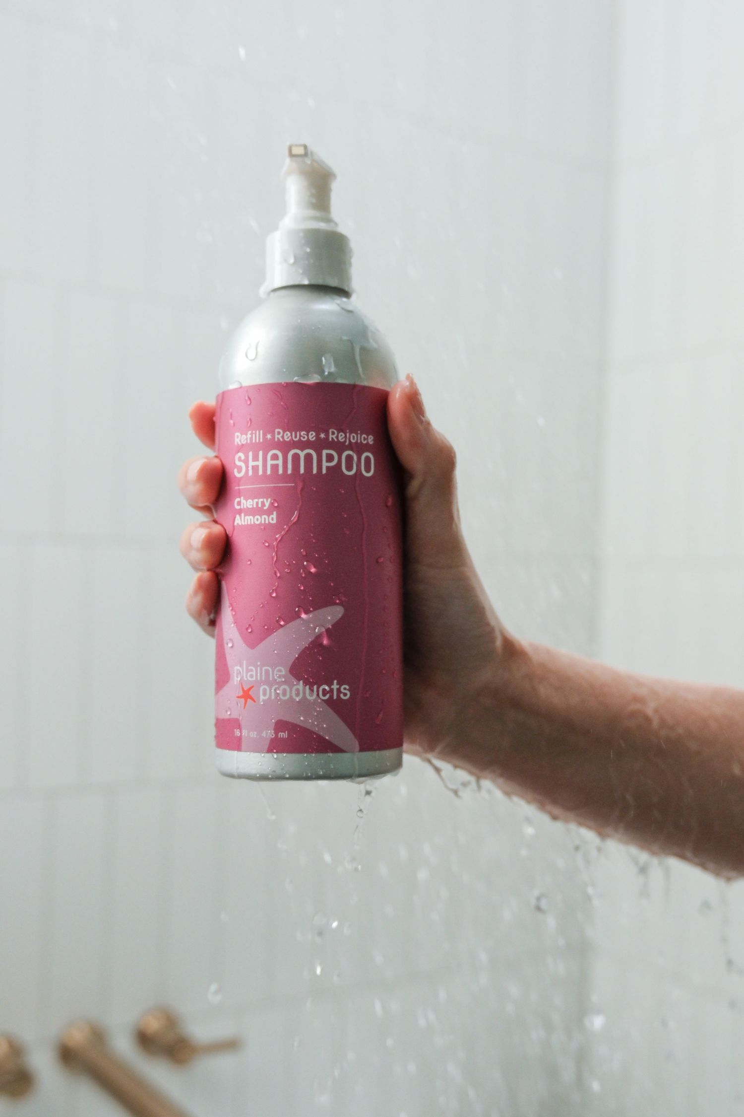 Hand holding Plain Products Cherry Almond Shampoo bottle with pink label in shower.