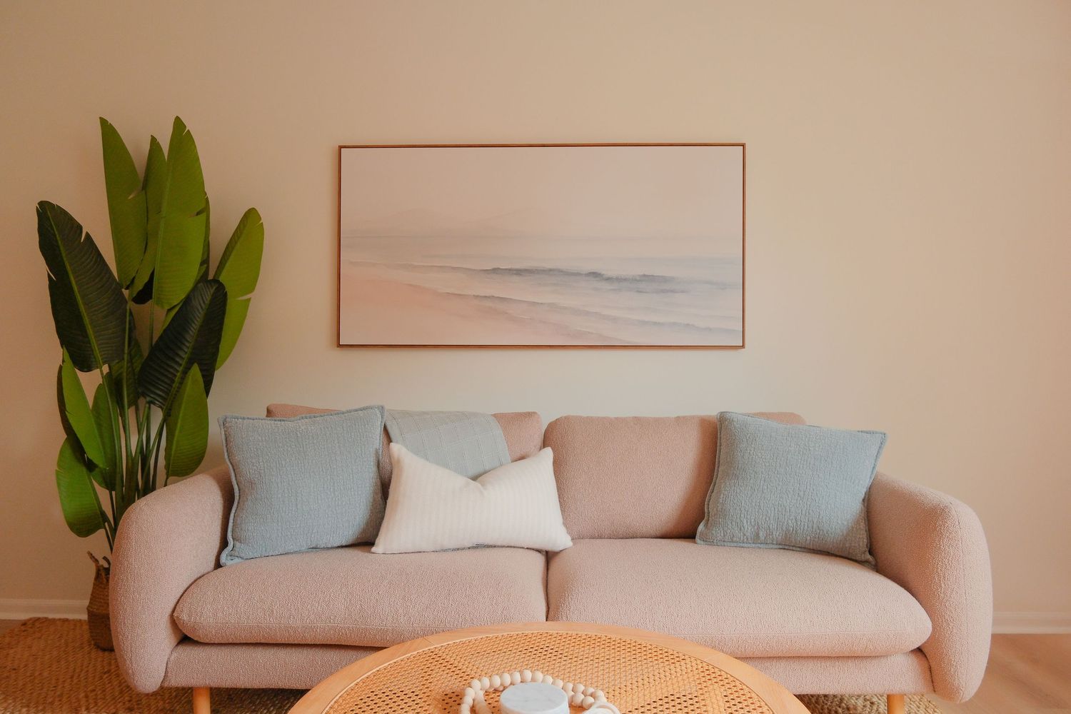Minimalist living room with a blush pink sofa, light blue pillows, coastal wall art, and a large green plant.