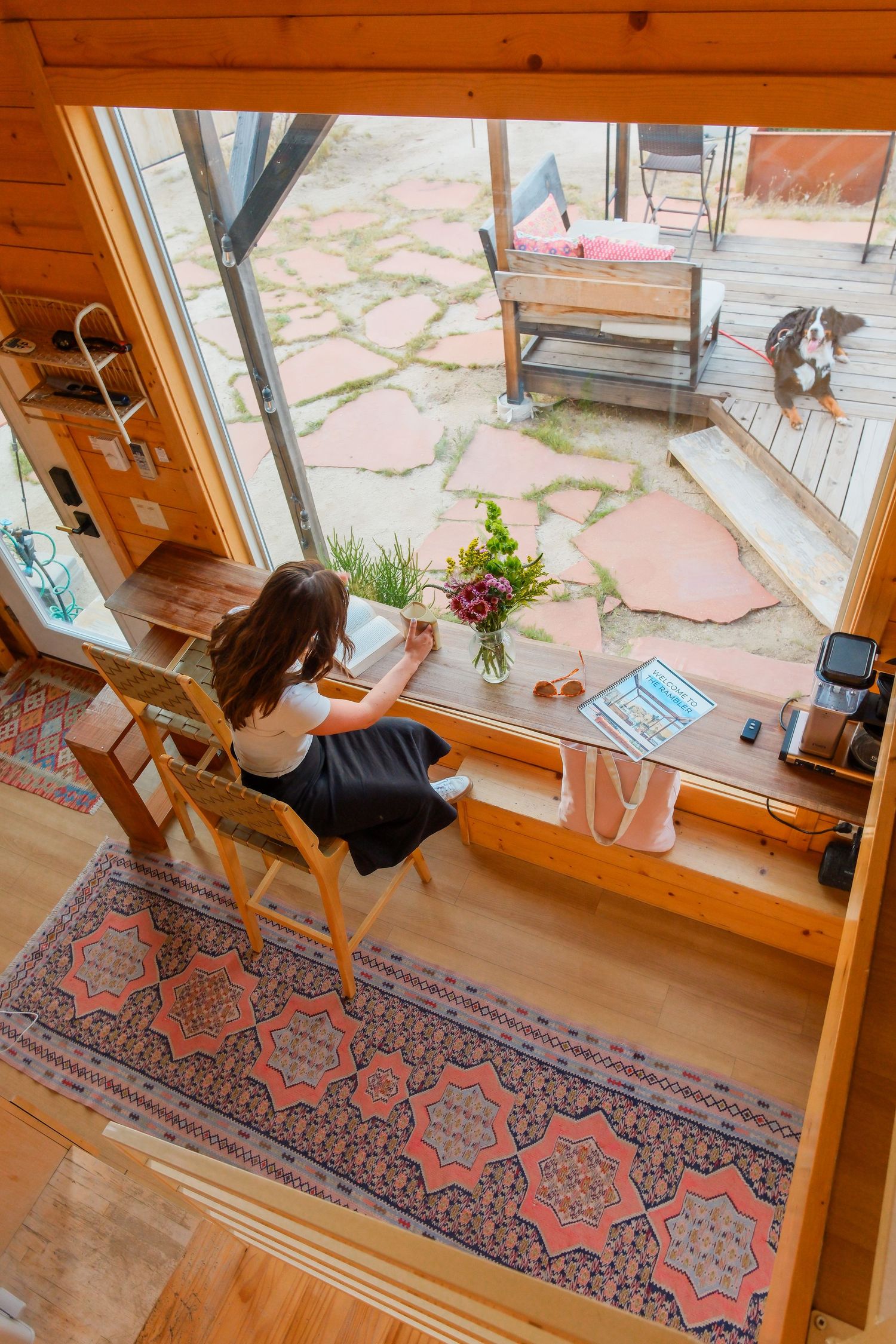 Aerial view of woman sitting at wooden desk by large window overlooking stone patio with dog in cozy cabin interior.