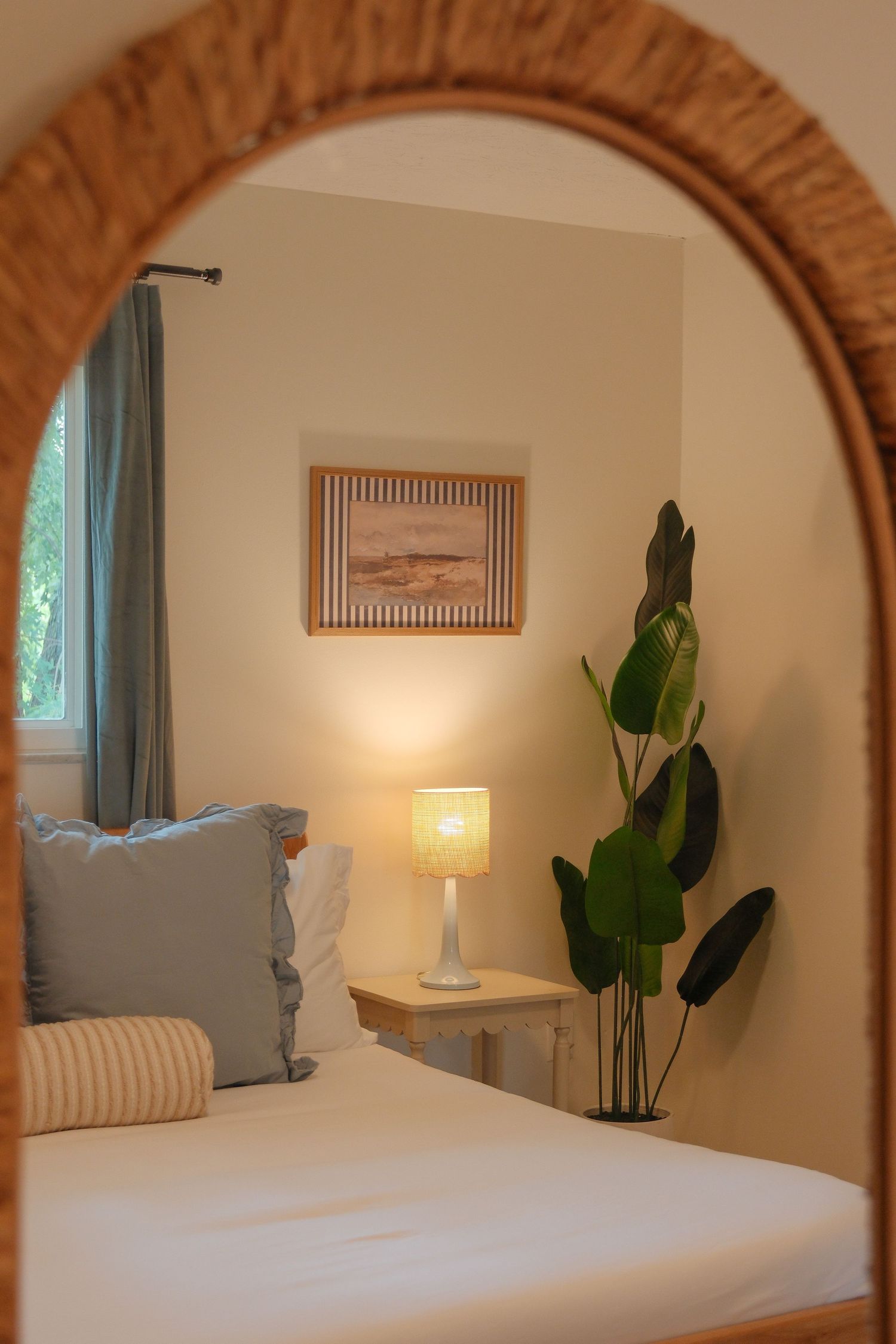 Cozy bedroom reflected in arched rattan mirror with glowing lamp, tropical plant, framed art, and soft blue pillows.