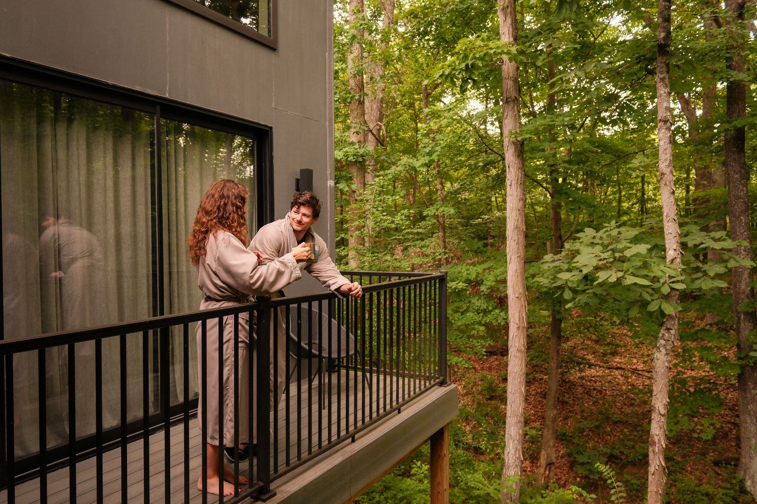 Couple in robes enjoying a wooded view from a modern cabin balcony surrounded by lush green trees.