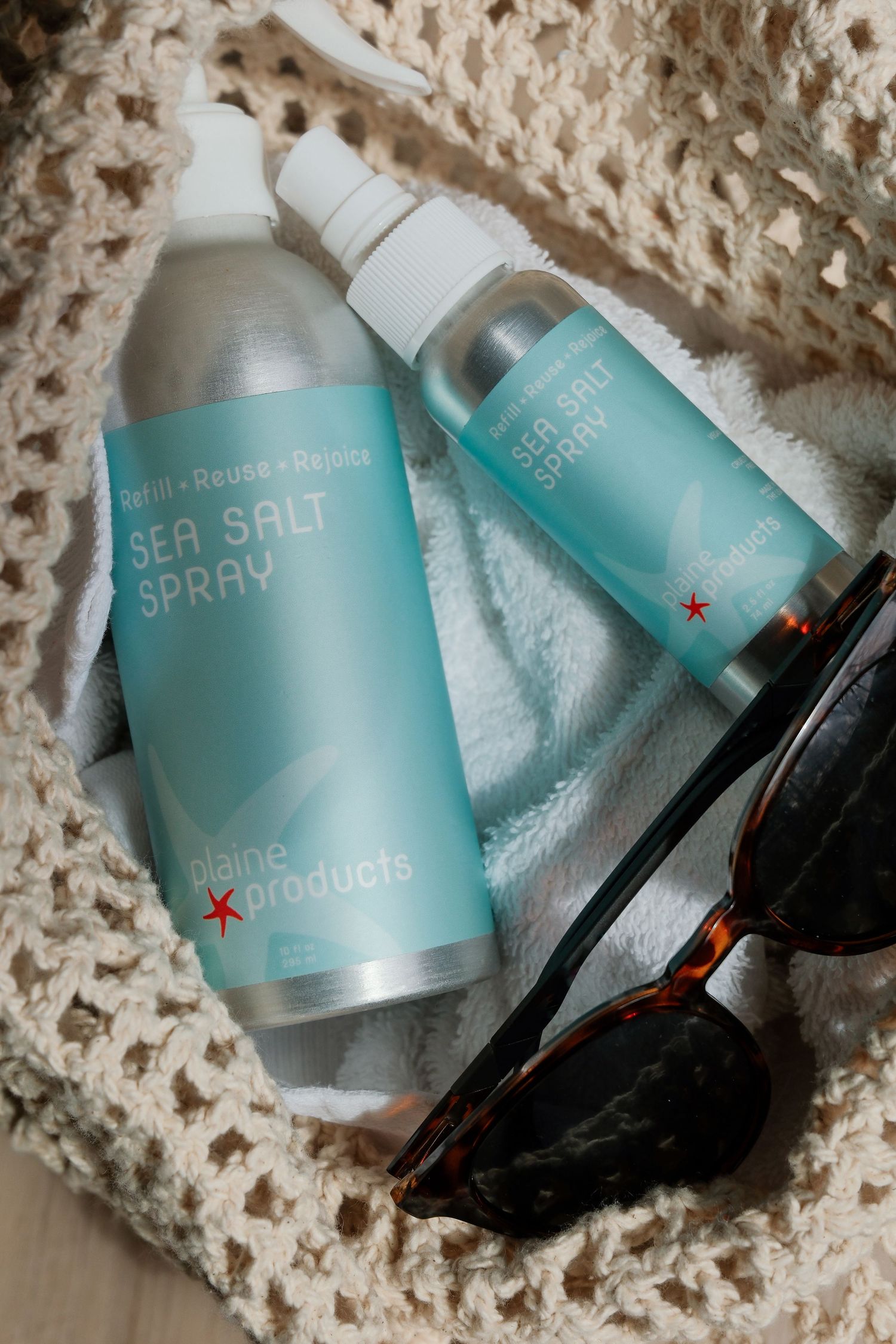 Two Plaine Products Sea Salt Spray bottles in a crochet bag with sunglasses on a white towel.