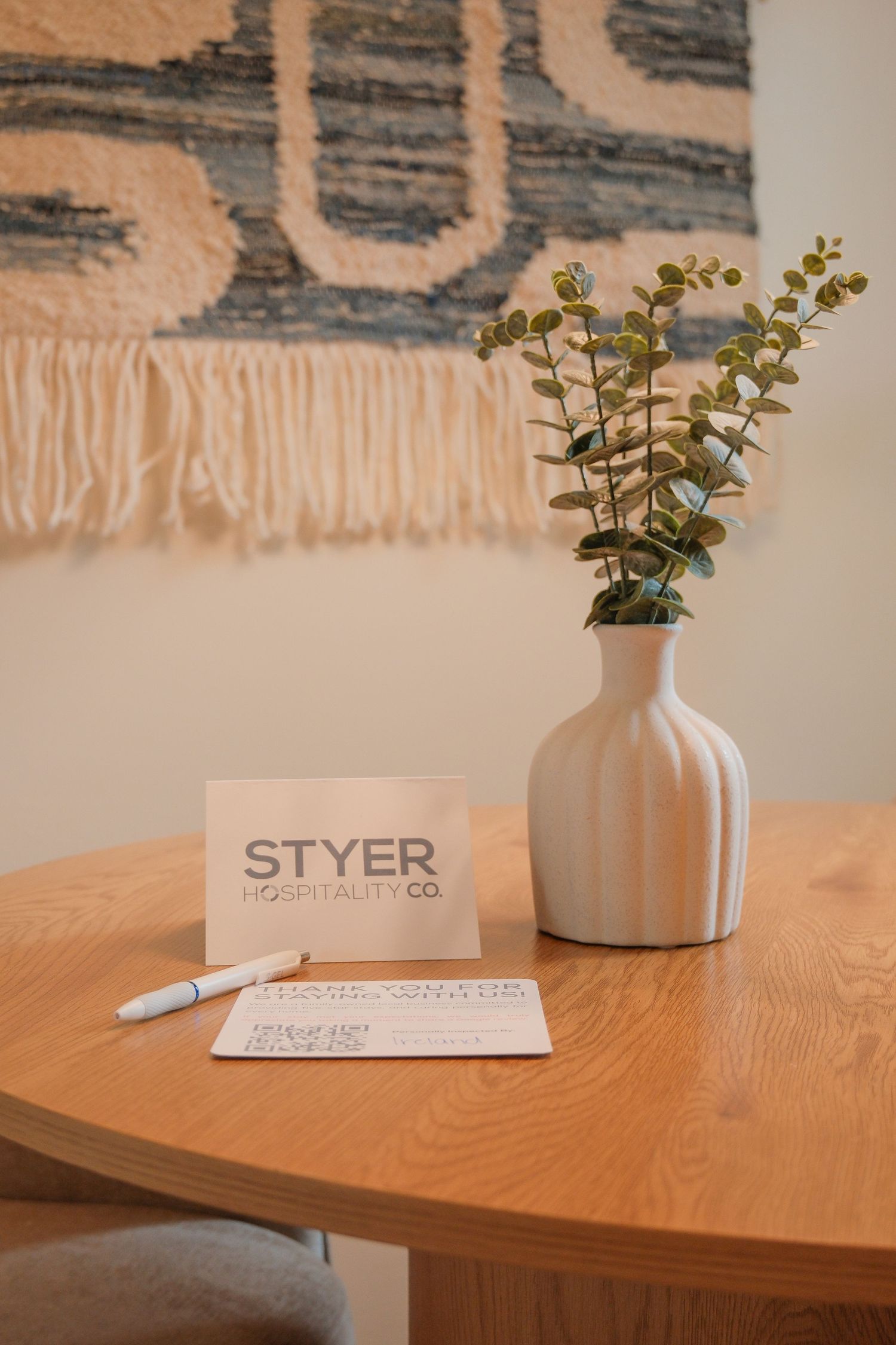 Styer Hospitality Co. branded card and pen on a wooden table with a ceramic vase holding eucalyptus stems.