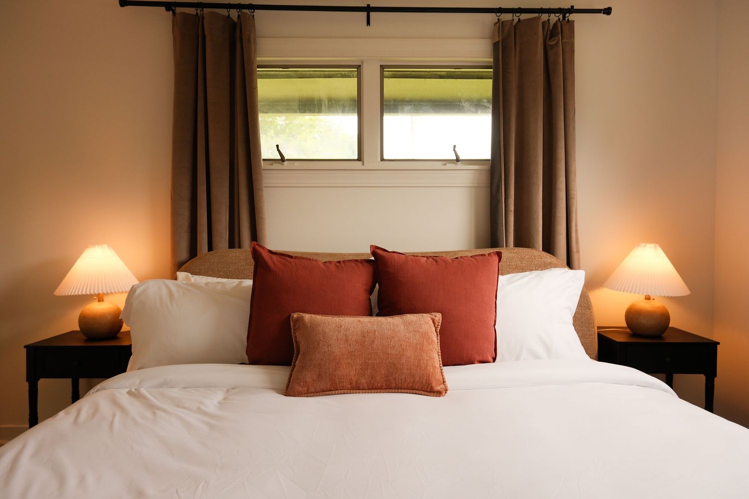 Cozy bedroom with white bedding, red and terracotta pillows, warm lamp lighting, and brown curtains framing a window.