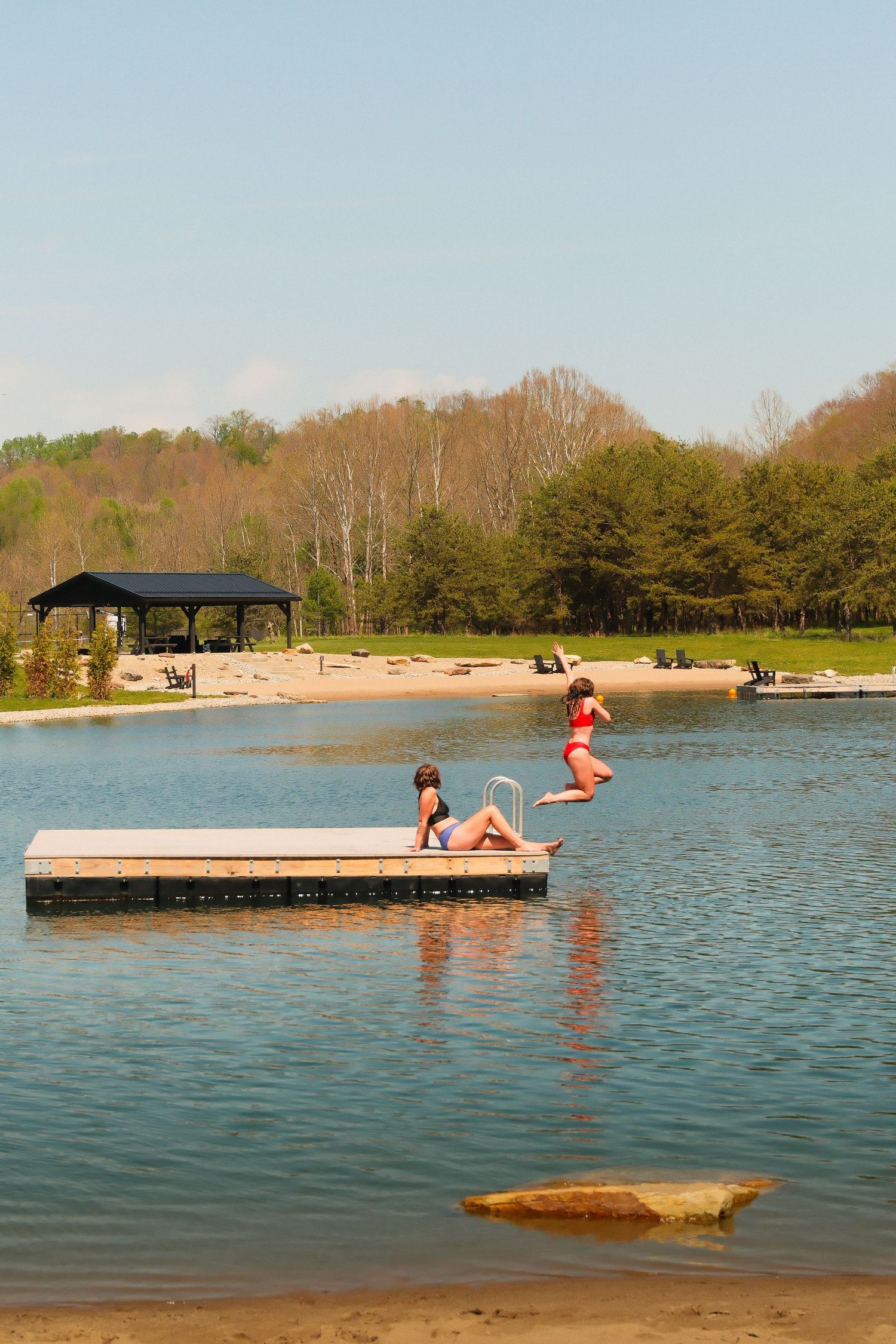 Two girls in swimsuits enjoy a sunny day on a floating dock at a calm lake with a sandy beach and pavilion.