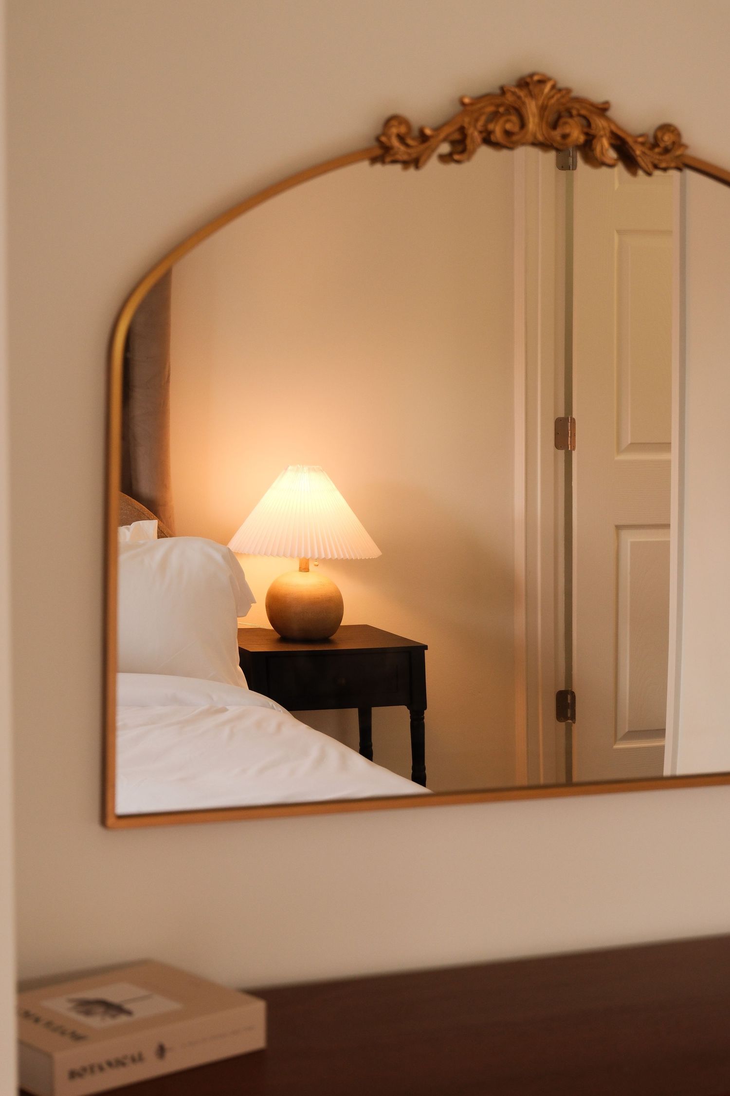Ornate gold arched mirror reflecting a cozy bedroom with a glowing bedside lamp on a dark nightstand.