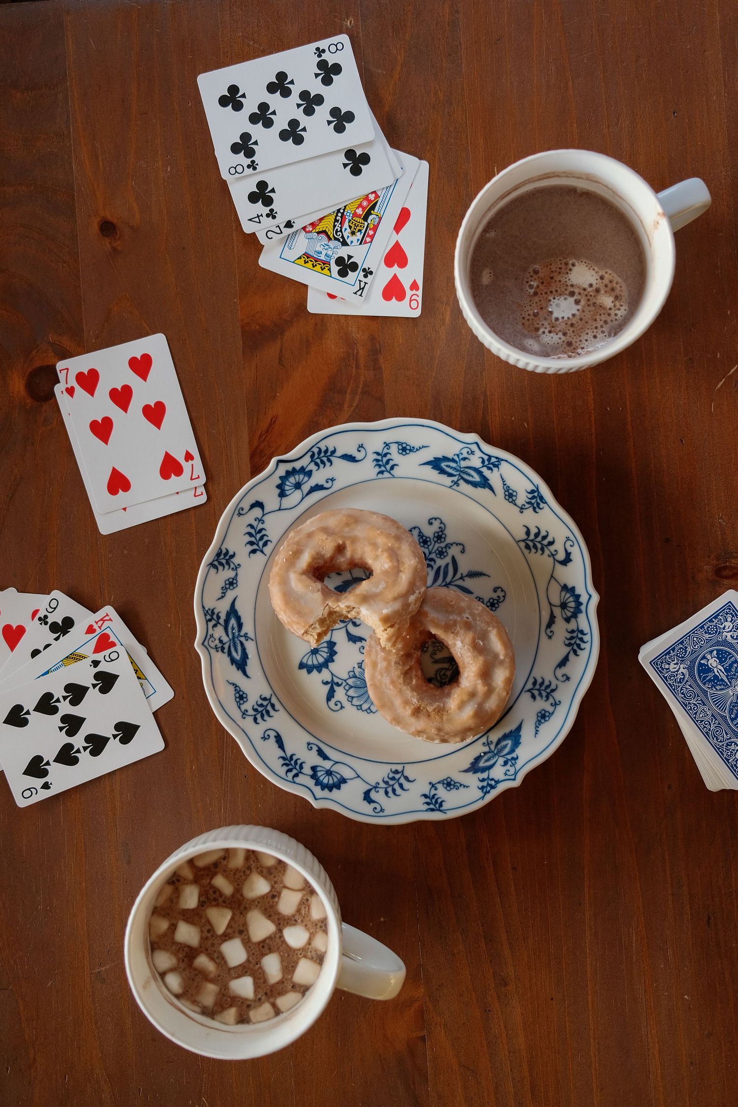 Playing cards scattered around hot chocolate and donuts on a wooden table with blue and white china.