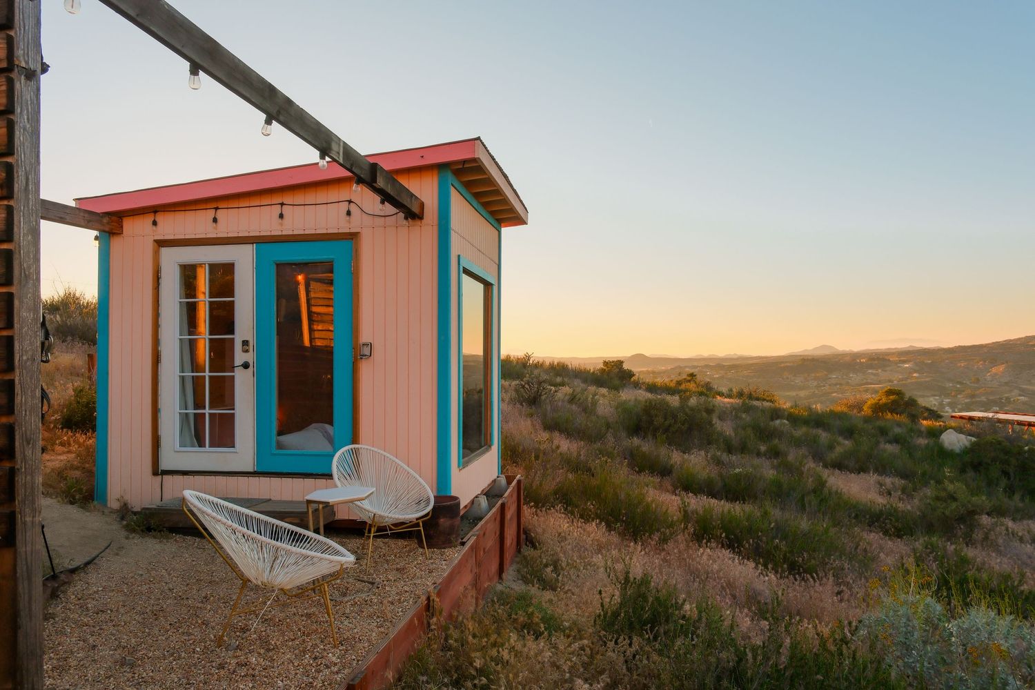 Tiny colorful cabin with turquoise door overlooking scenic desert valley at golden sunset.