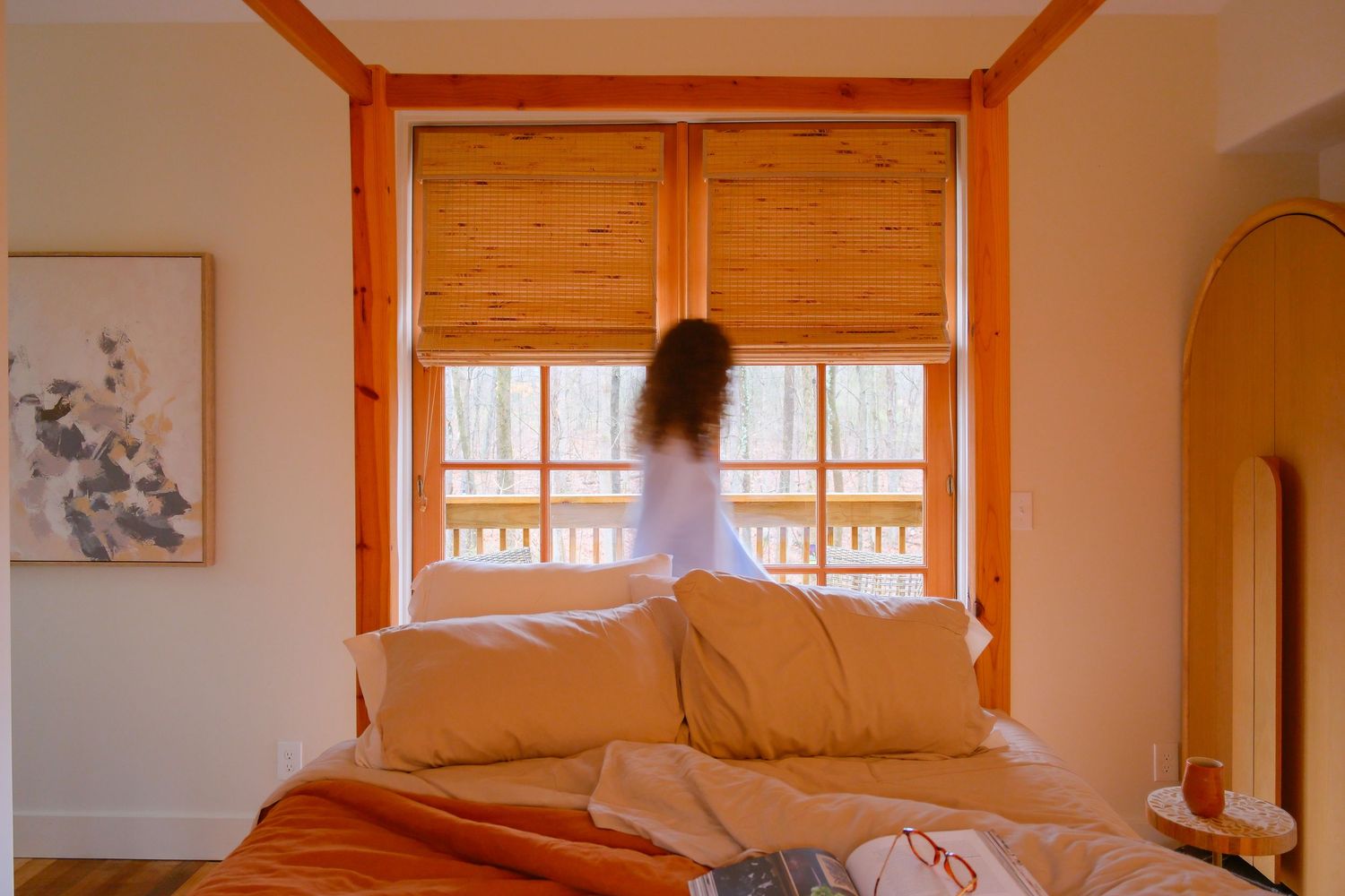 Woman standing at bamboo shade windows in a cozy bedroom with orange bedding and warm wood accents.