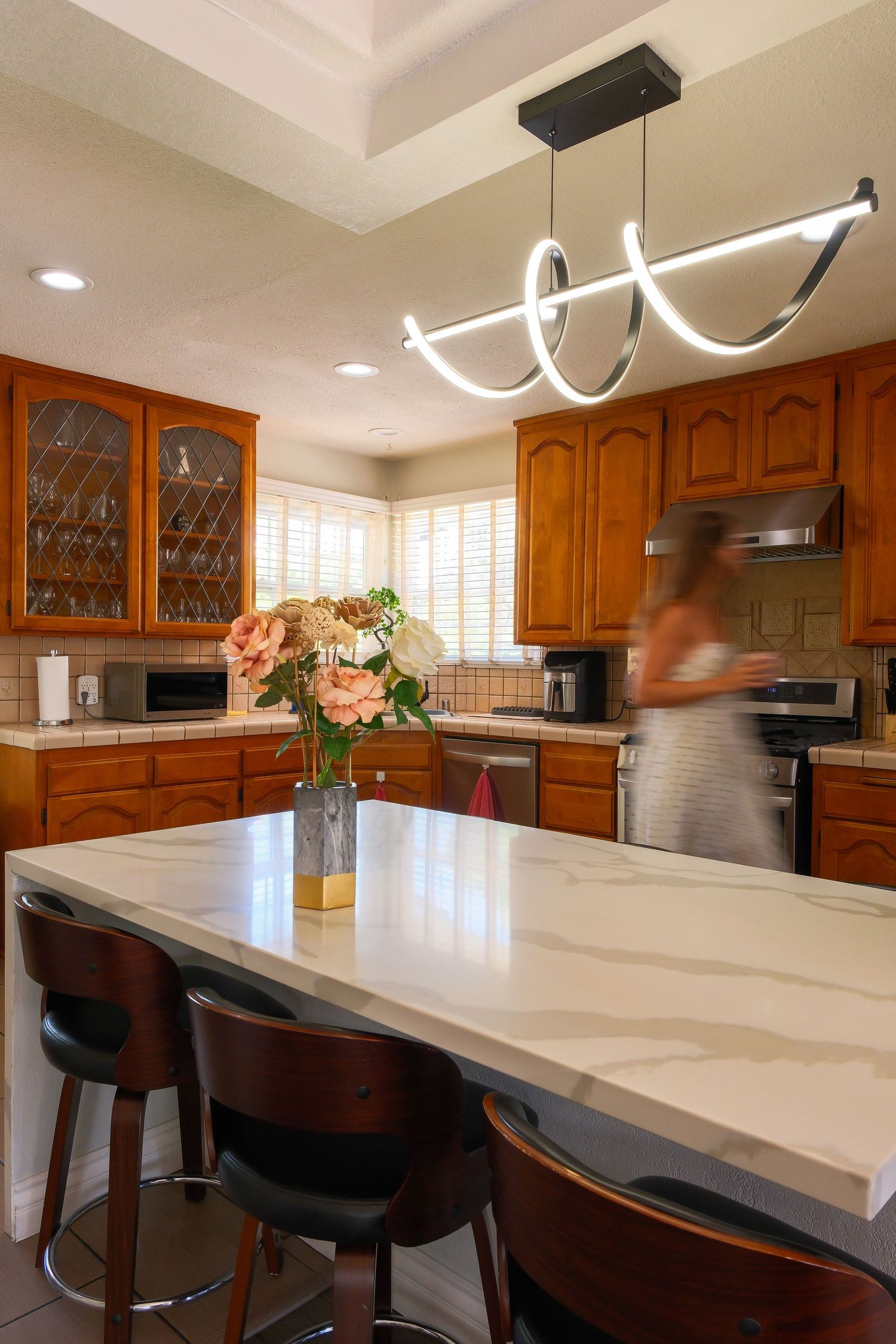 Modern kitchen with warm wood cabinets, marble island, bar stools, and contemporary LED pendant light fixture.
