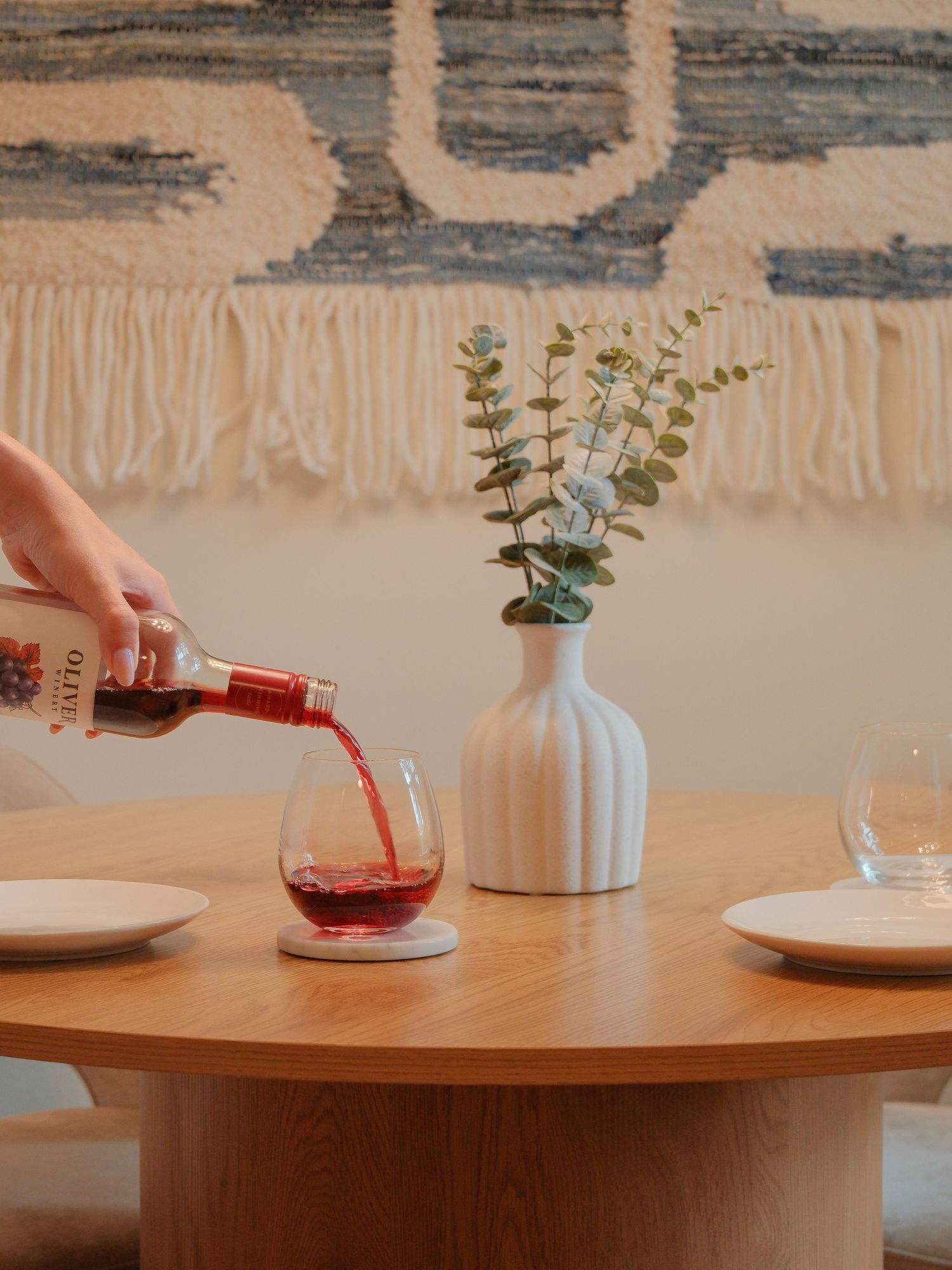 A hand pouring red wine into a stemless glass on a wooden round table with a white vase of eucalyptus and boho wall tapestry.