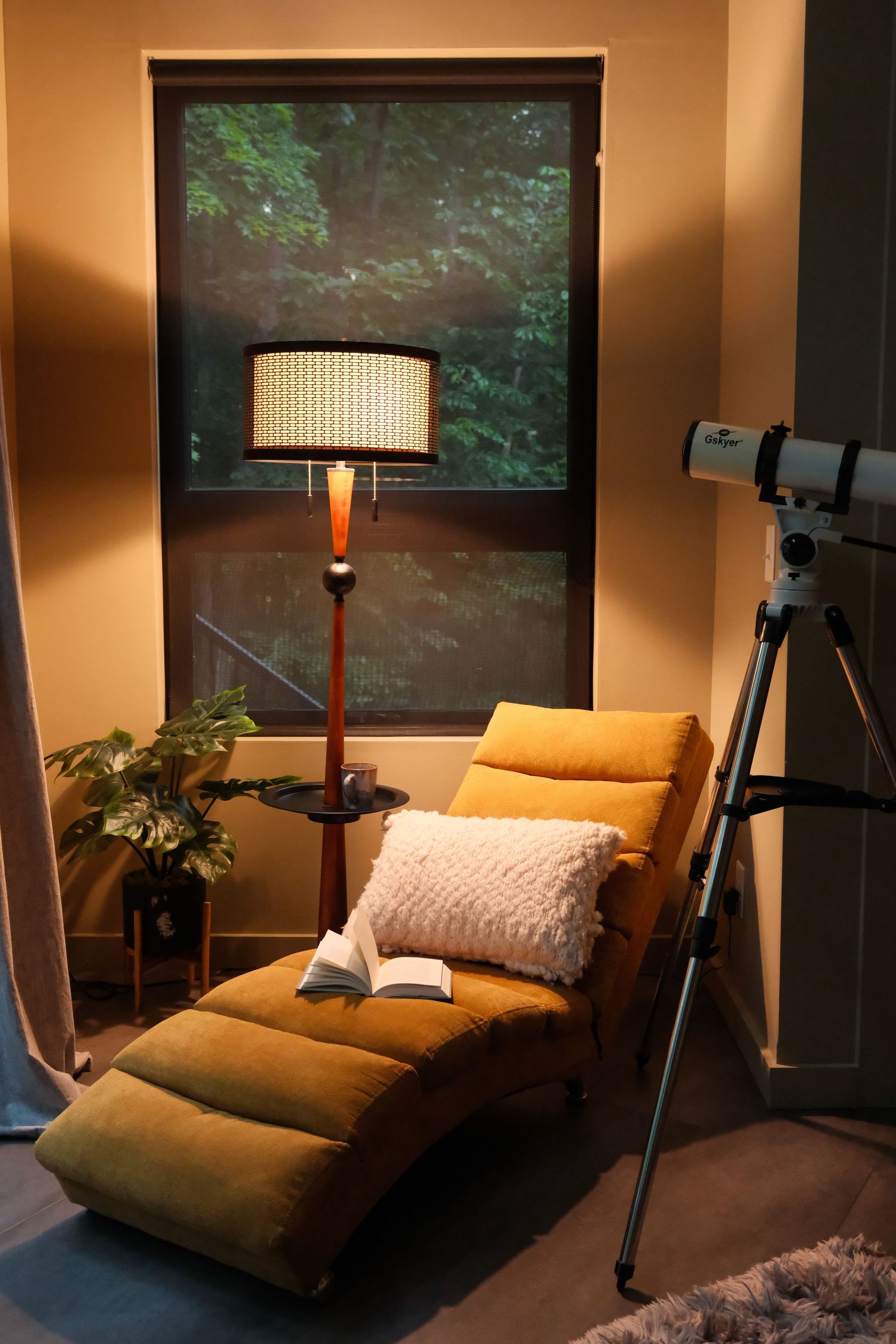 Cozy reading nook with mustard yellow chaise lounge, floor lamp, open book, pillow, and telescope near a window at night.