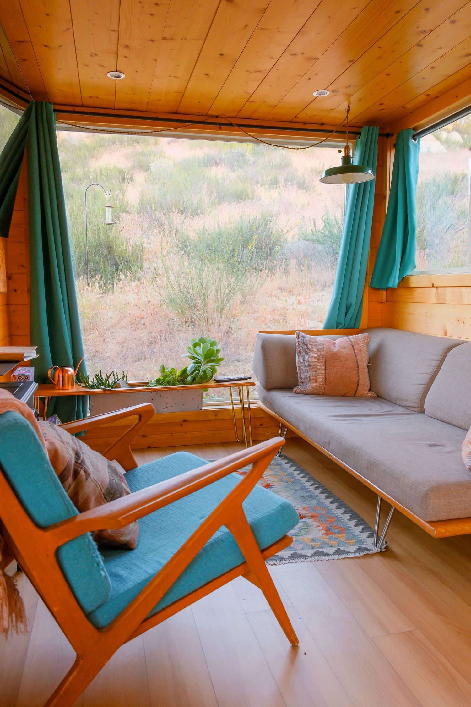 Cozy tiny home living room with teal curtains, mid-century modern chair, gray sofa, and wood paneling overlooking nature.