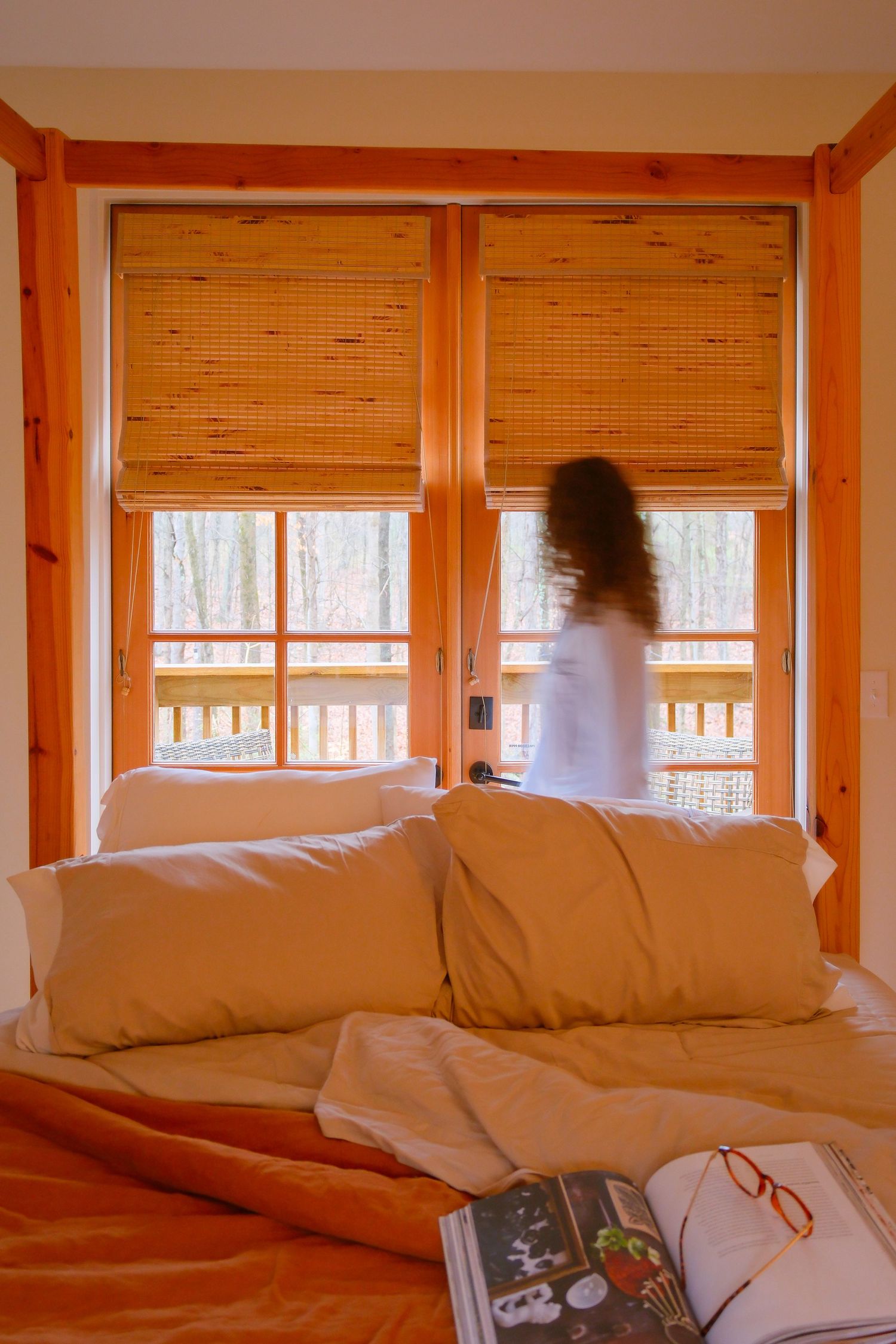 Cozy wooden cabin bedroom with orange bedding, bamboo blinds, open book, and blurred woman standing by glass door.