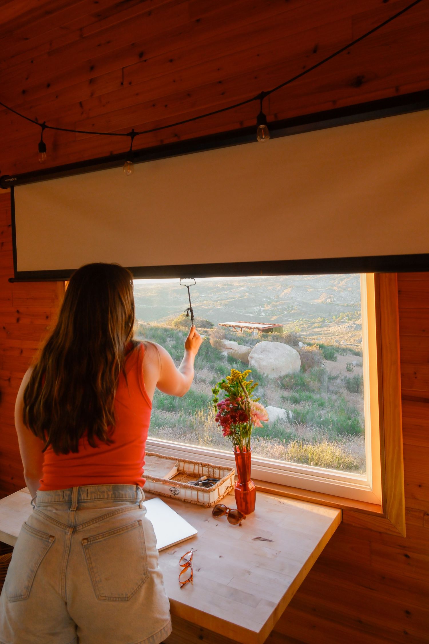 Woman in orange top adjusting projector screen above large window with scenic mountain landscape view.