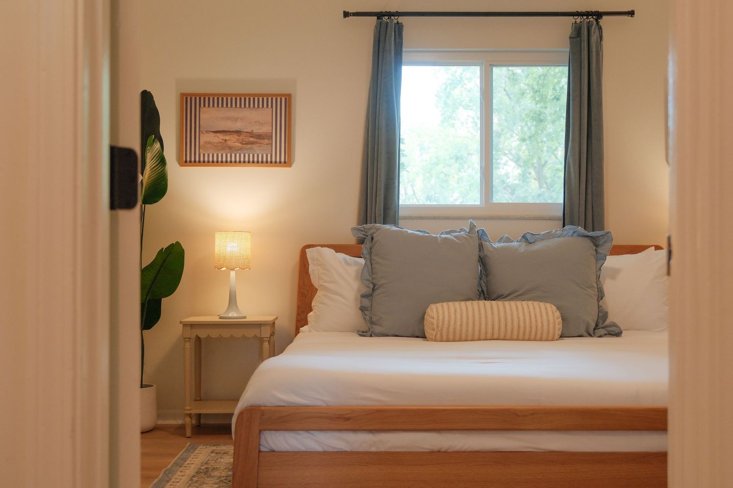 Cozy bedroom with wooden bed, gray pillows, bedside lamp, and window with teal curtains.