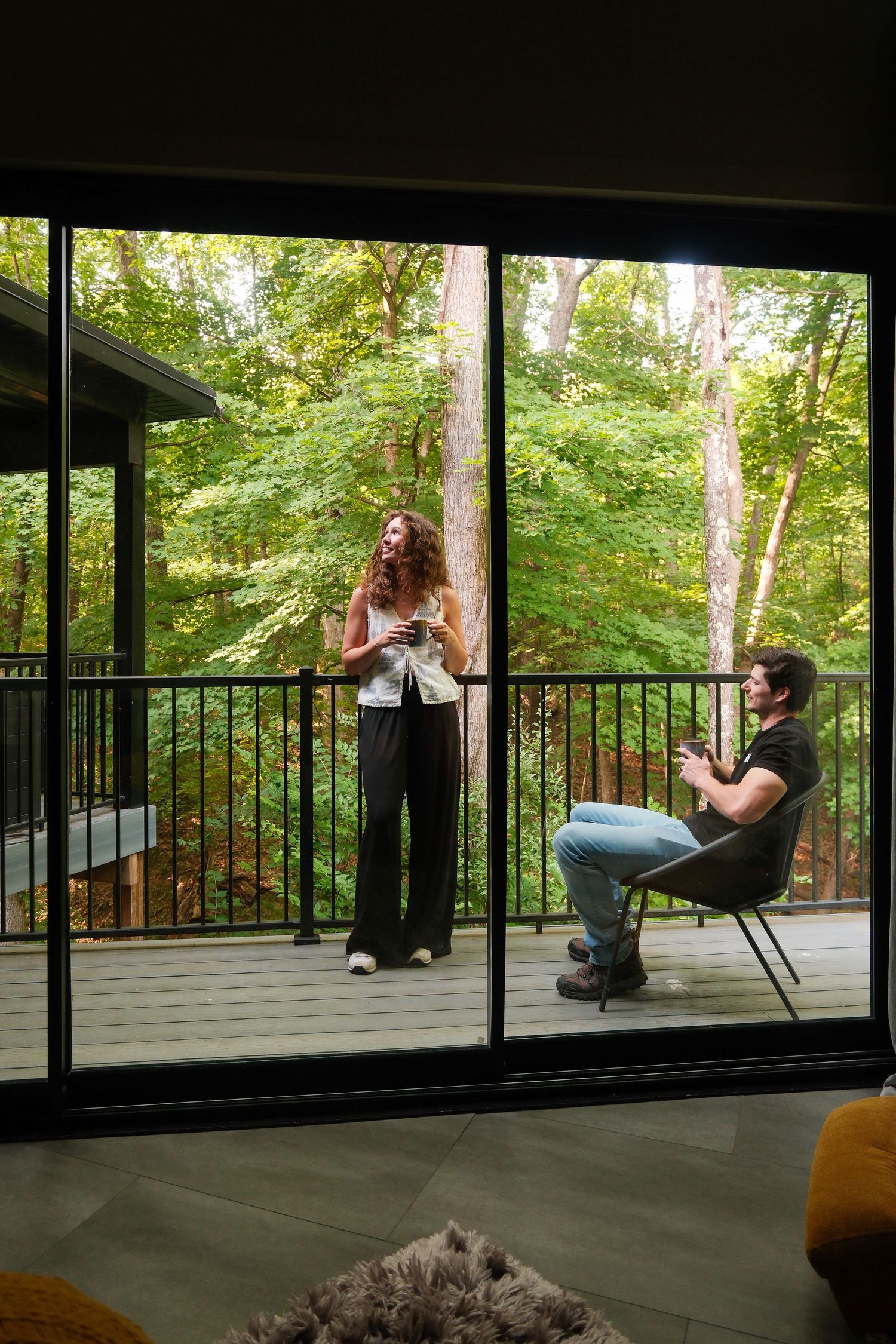 A couple enjoying drinks on a wooded deck, viewed through large sliding glass doors of a modern home.