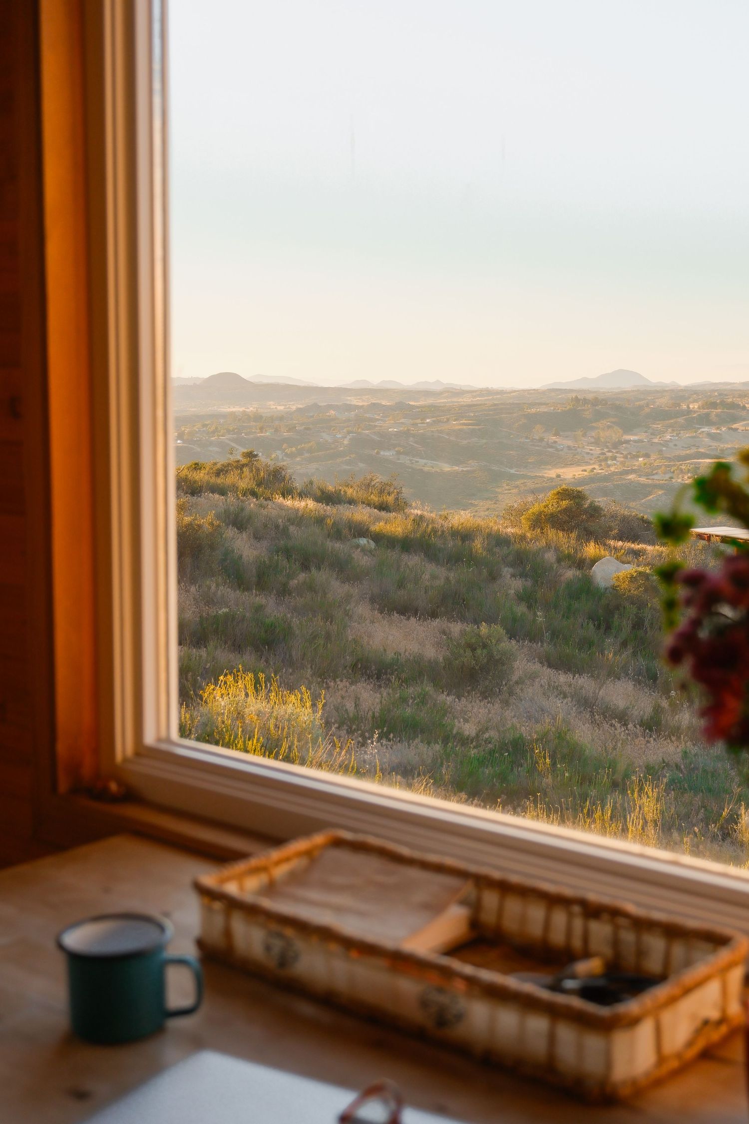 Cozy window view of rolling green hills at sunset with a teal mug and wooden tray on the sill.