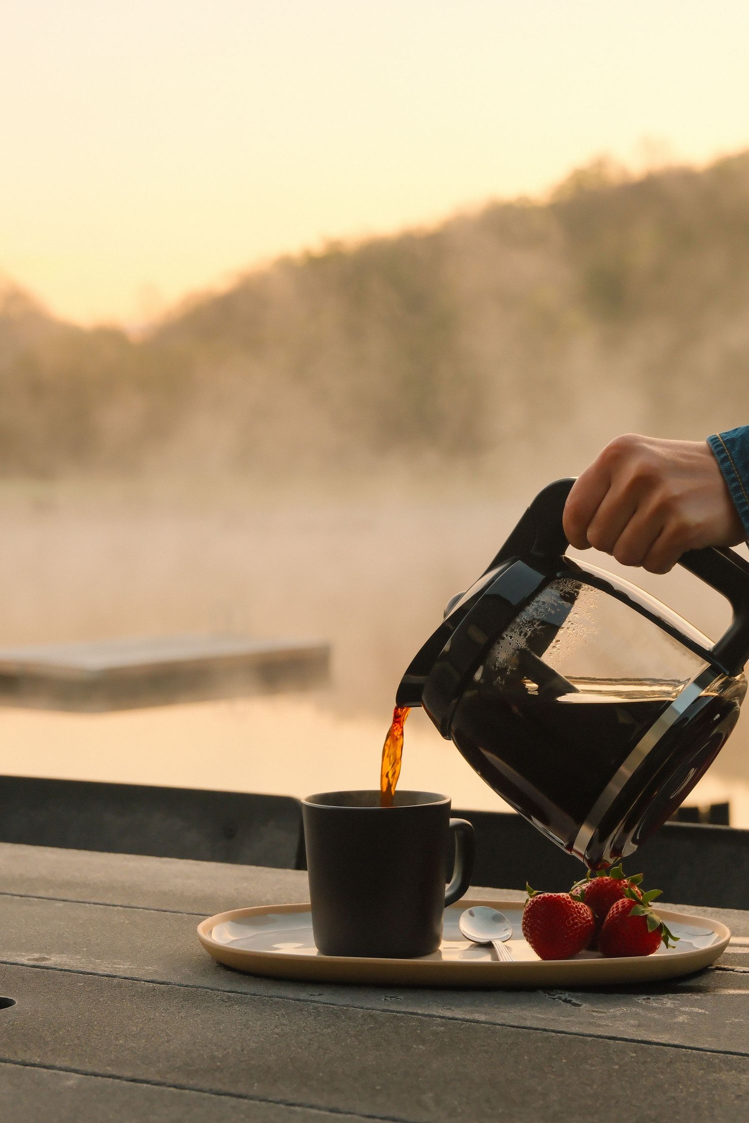 Hand pouring coffee into a dark mug on a tray with strawberries, set against a misty lake sunrise backdrop.