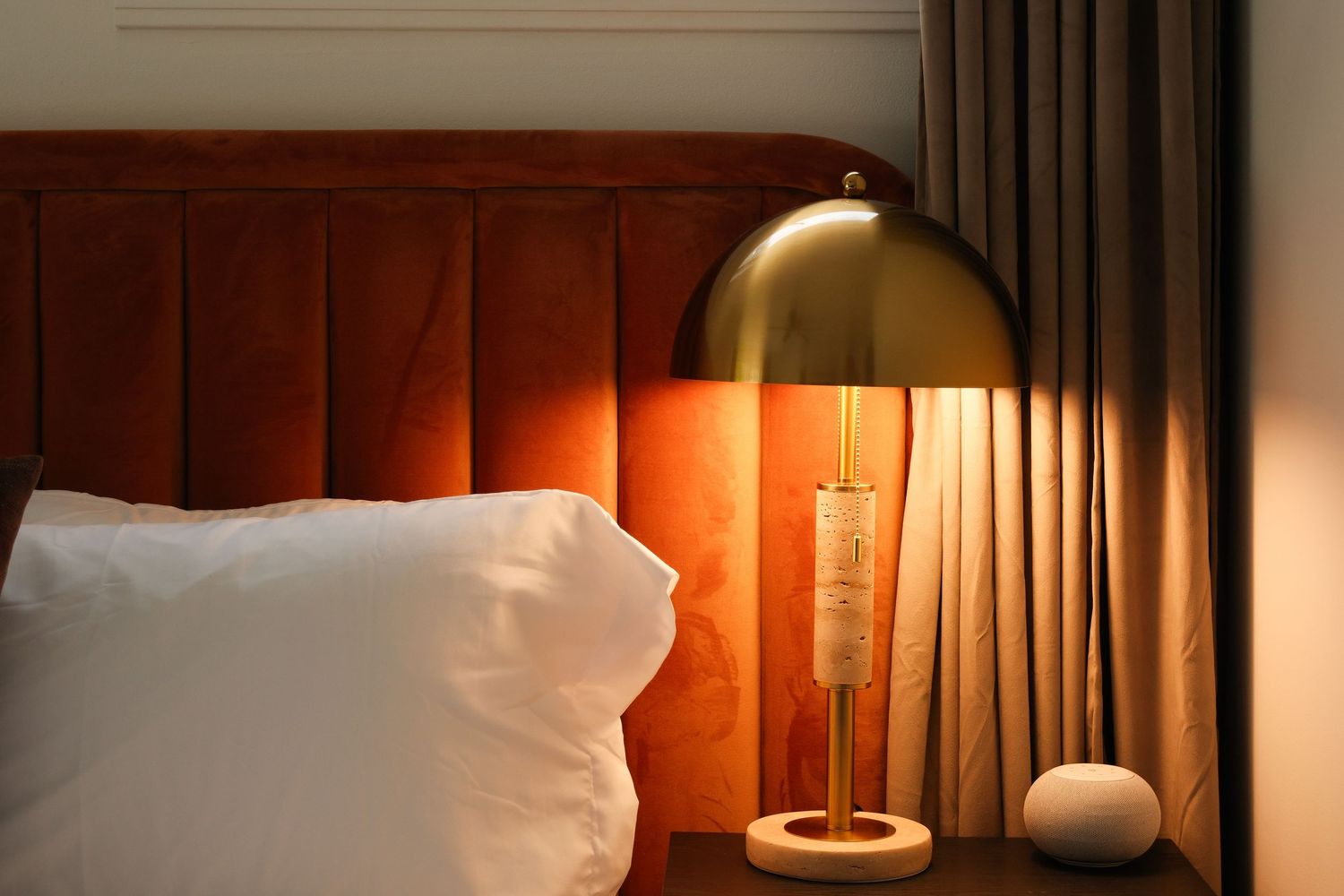 Elegant hotel bedroom with a gold dome table lamp on a marble nightstand beside a velvet orange headboard.