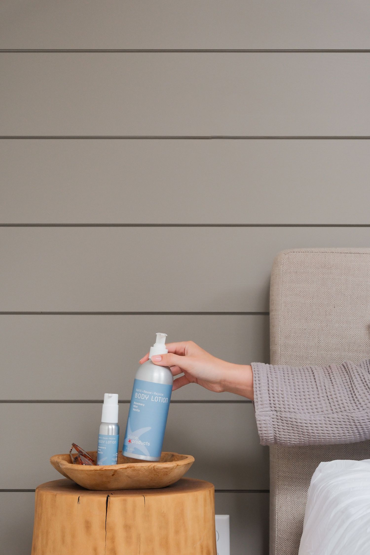 Hand reaching for large blue spray bottle on wooden stump nightstand beside gray upholstered headboard.