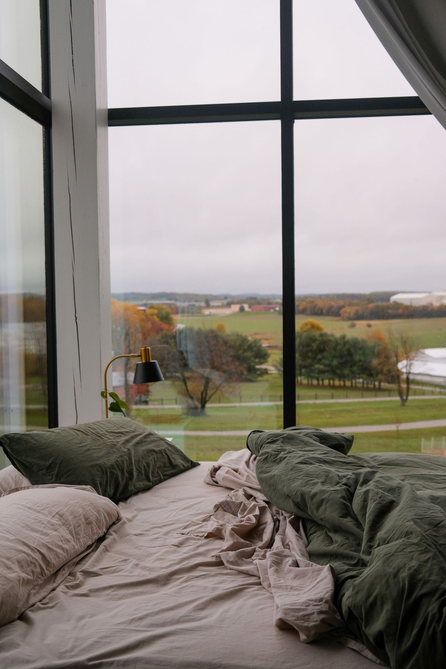 Unmade bed with green and beige linens beside large windows overlooking autumn countryside.