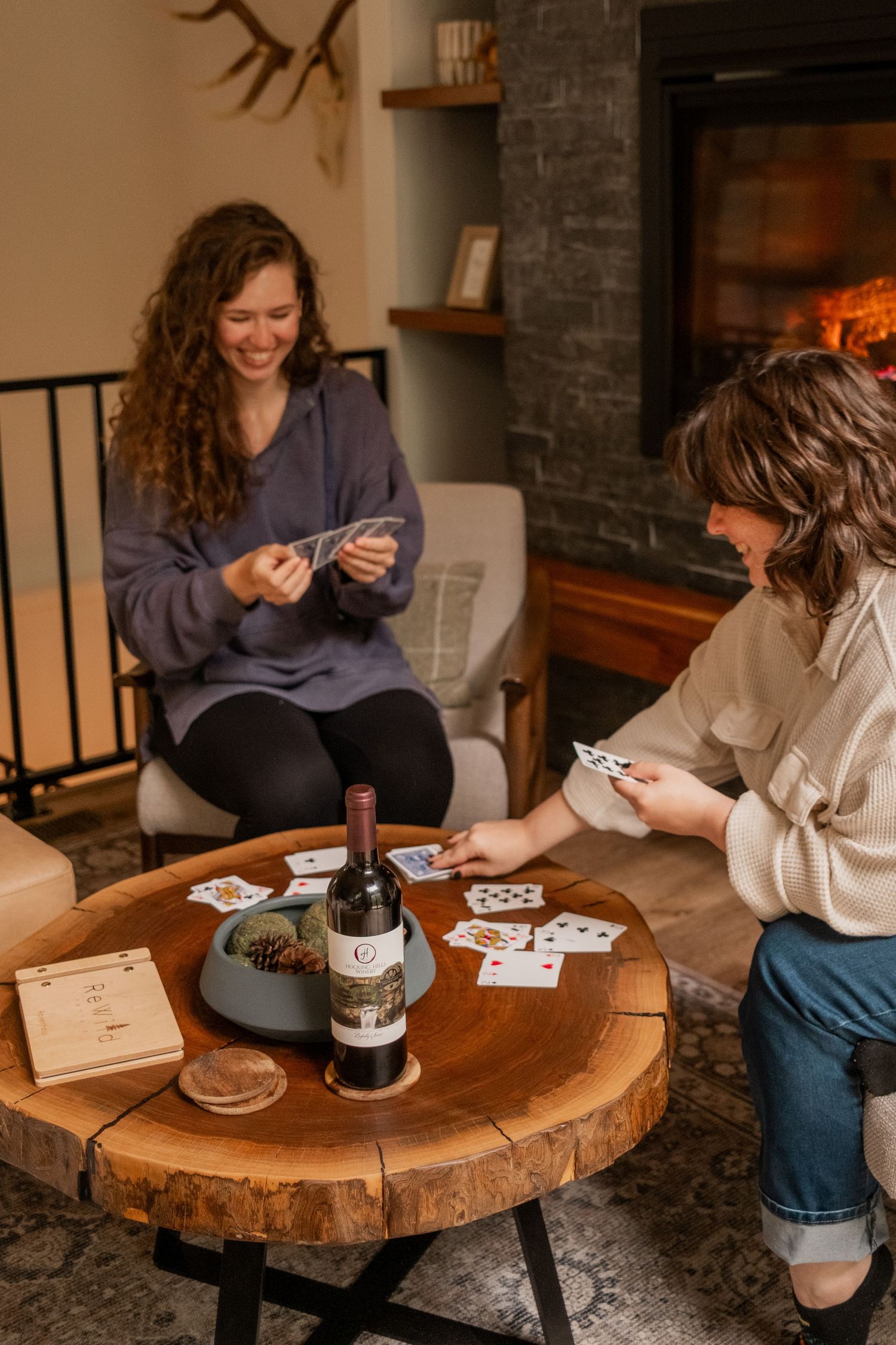 Two people playing cards on a rustic wood table with wine and cozy lighting in the background.